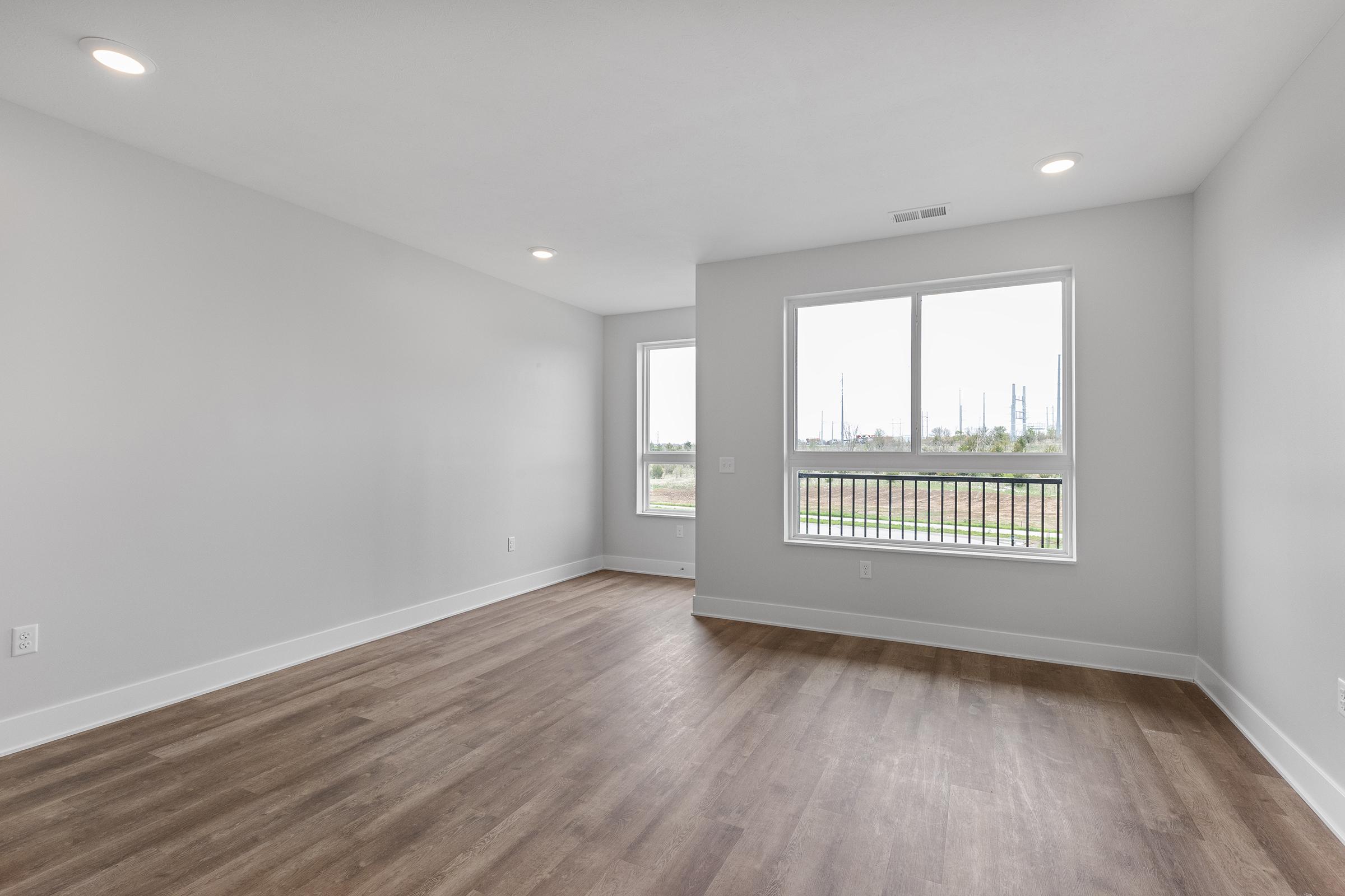 A spacious, empty room with light gray walls and hardwood flooring. It features large windows on one wall, allowing natural light to flood in, and a small balcony. The view outside shows greenery and industrial structures in the distance. The overall atmosphere is bright and airy.