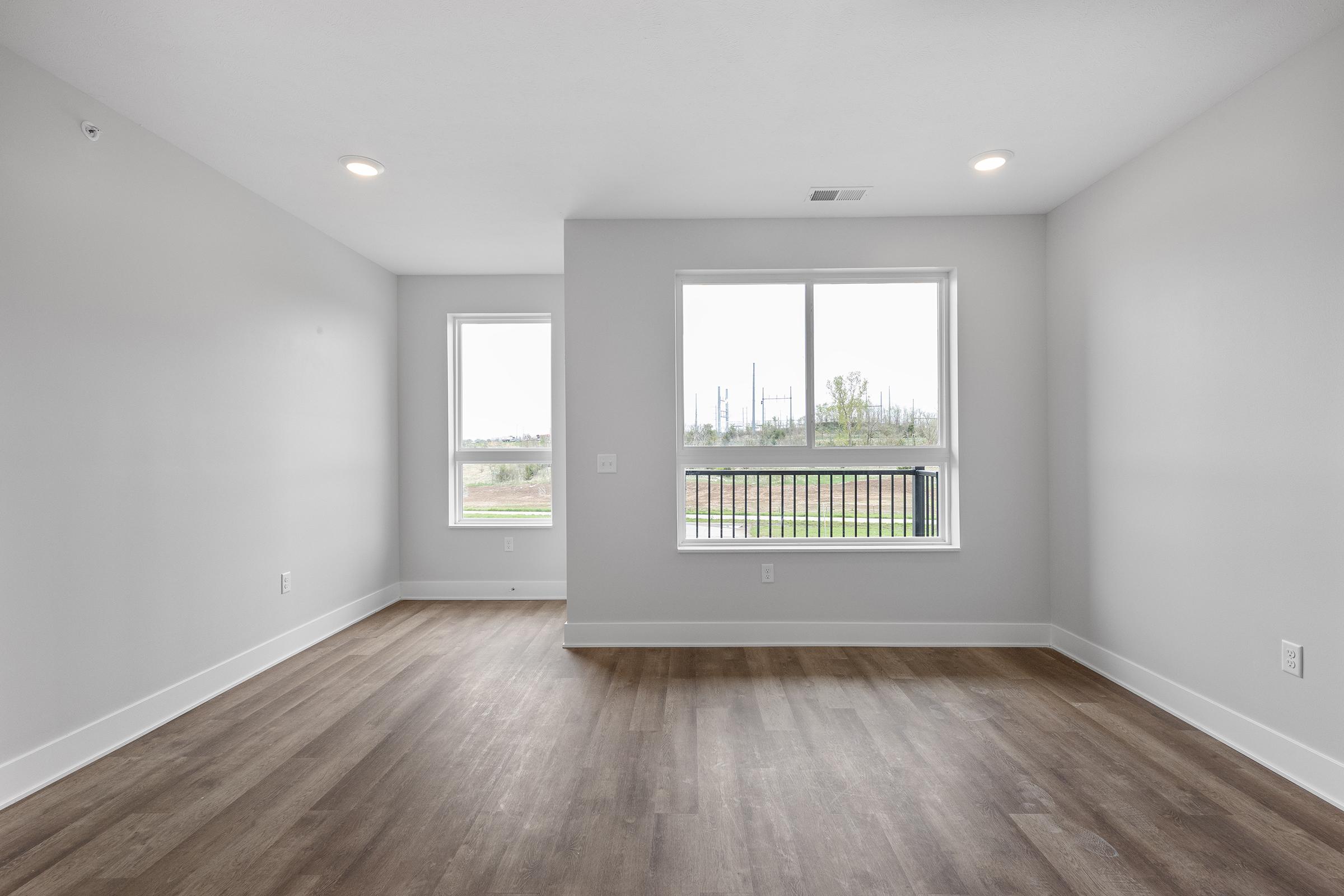 A spacious, empty room with light gray walls and new hardwood flooring. Two windows provide natural light, showcasing a view of greenery outside. The space is bright and inviting, ideal for personalization and decoration.
