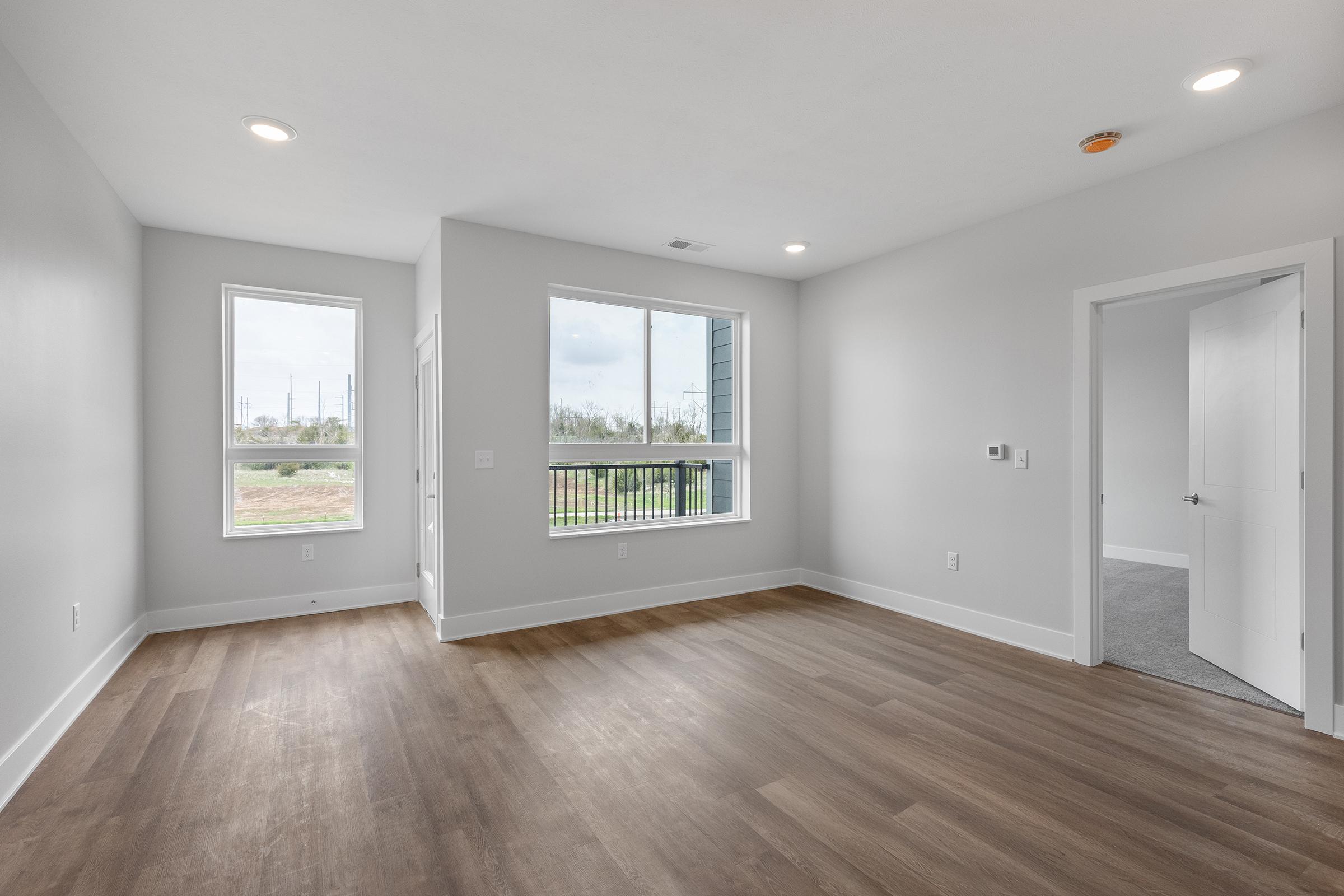 A spacious room with light gray walls and hardwood flooring. It features two windows allowing natural light, a door leading outside, and another door that opens to a hallway. The room is empty, providing a blank canvas for decoration.