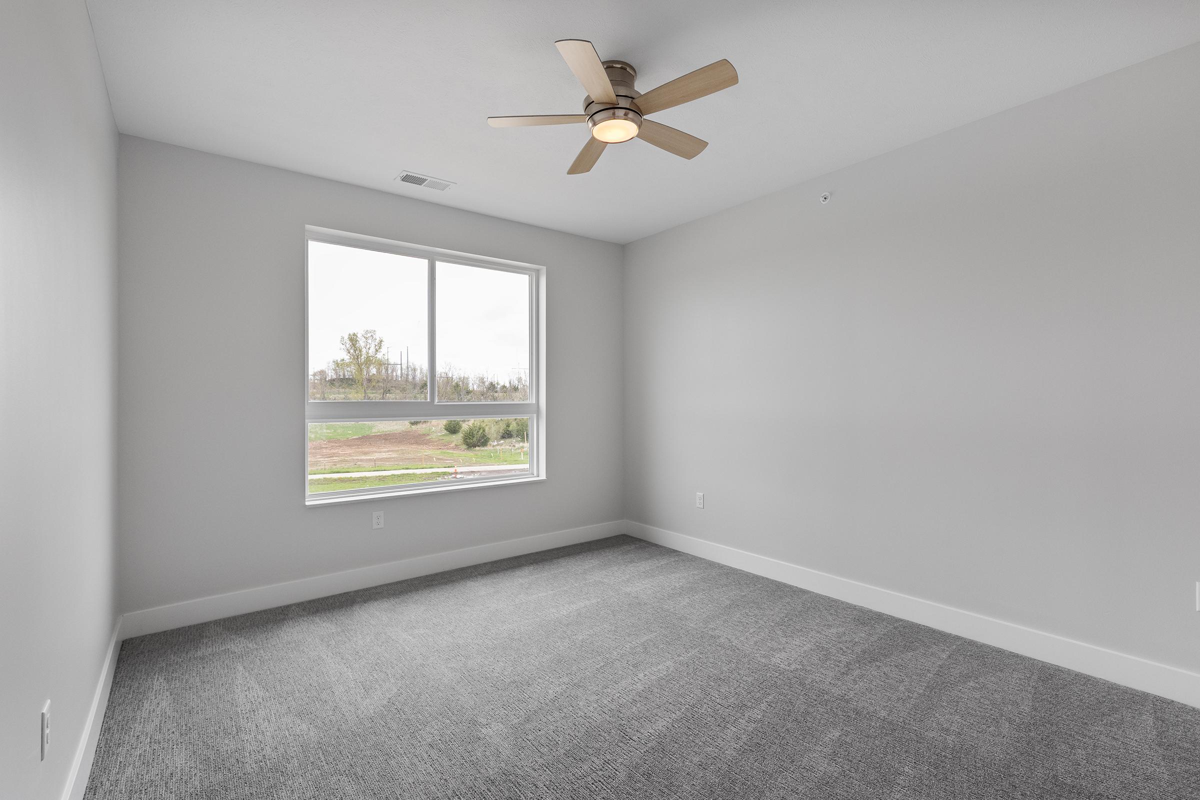 Empty room with light gray walls and carpet, featuring a ceiling fan. A large window lets in natural light, revealing a view of greenery outside. The space is minimally decorated, creating a clean and modern atmosphere.