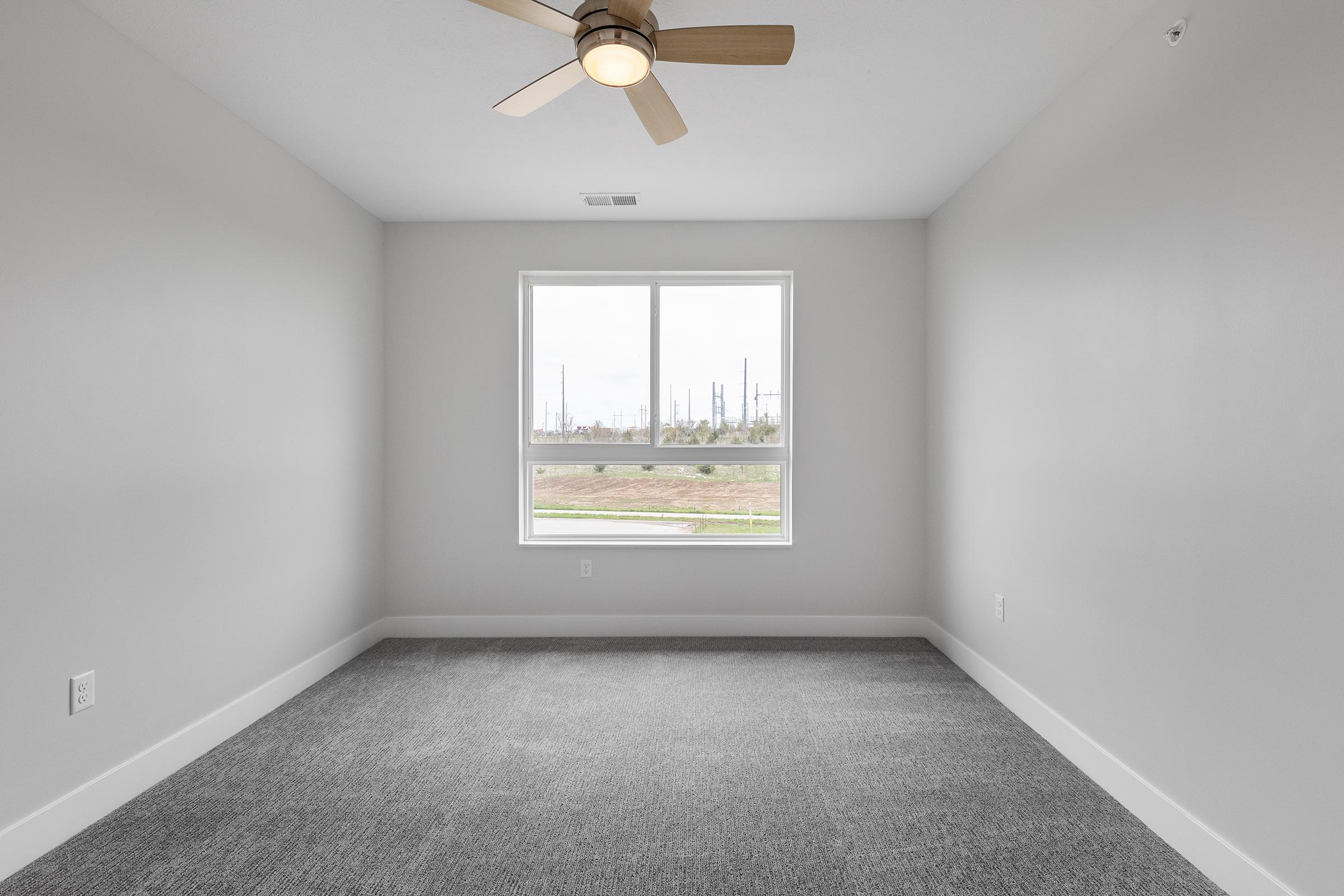 Empty room with gray walls and carpet, featuring a ceiling fan and a large window that offers a view of an outdoor area, possibly with industrial structures in the background. Natural light fills the space, making it feel open and airy.