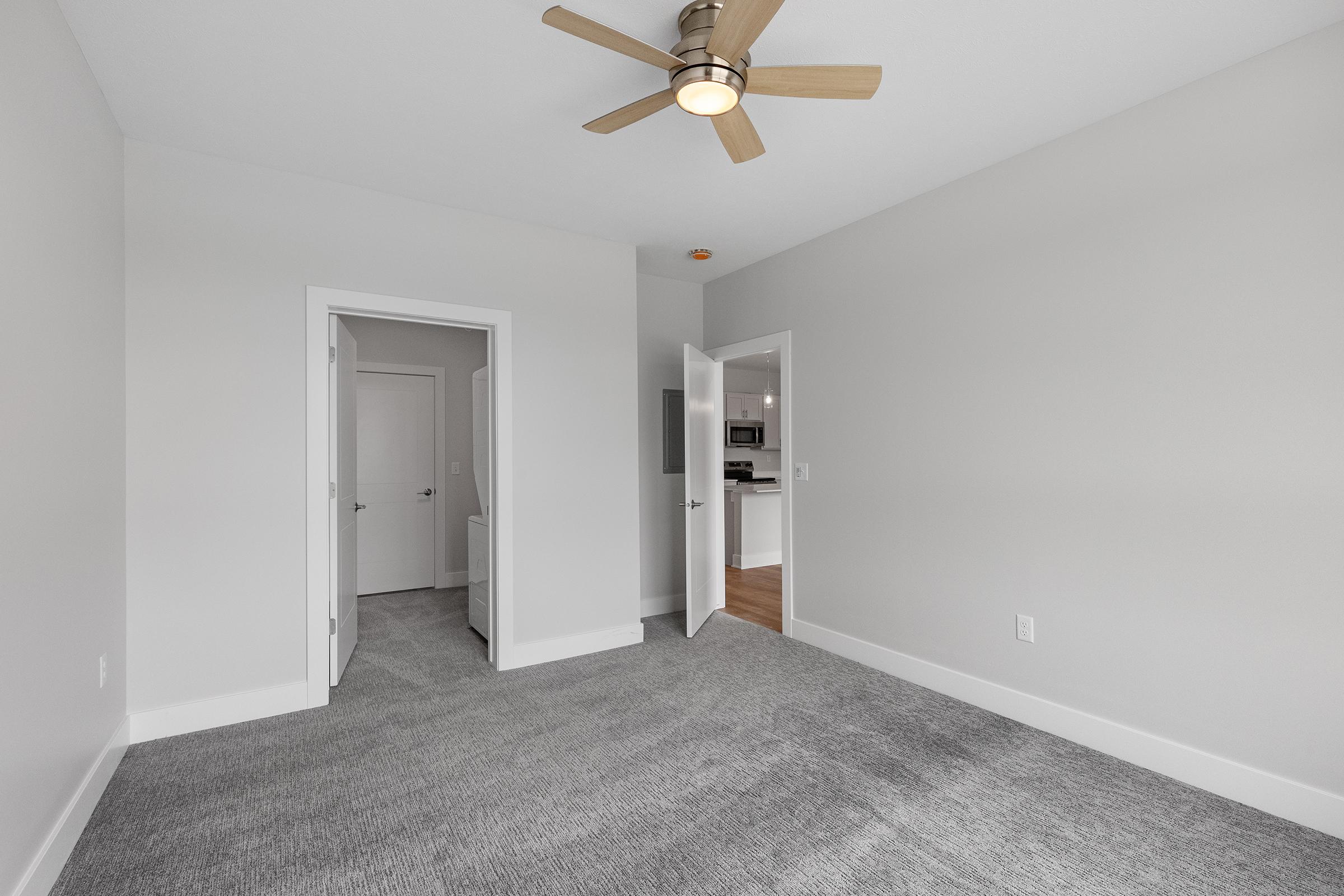 A spacious, well-lit room with light gray walls and plush gray carpeting. A ceiling fan is mounted on the white ceiling. Two doors lead to other areas, one opening to a kitchen or living space. Natural light brightens the room, creating a cozy atmosphere.