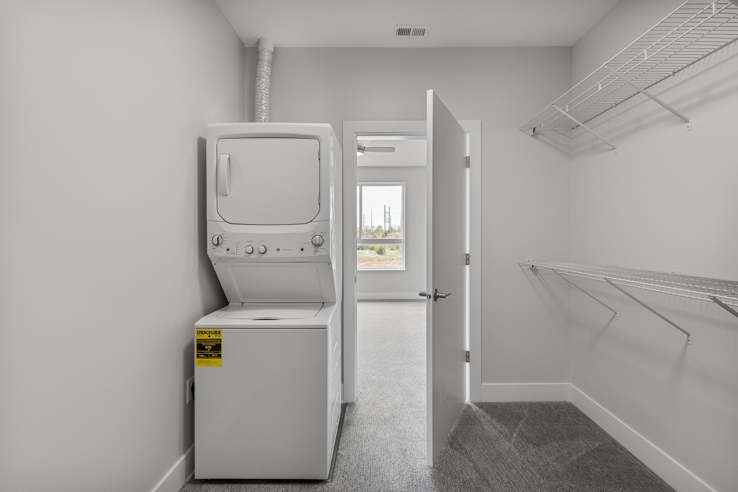 A modern laundry room featuring a stacked washer and dryer against a light gray wall. A door partially open leads to a bright room with a window showcasing a view of the outdoors. A wire storage shelf is mounted on one side, and the floor is carpeted in a soft gray.
