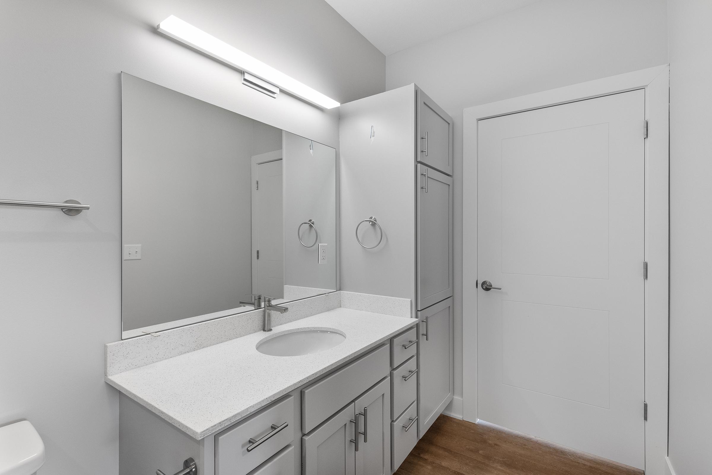 A modern bathroom interior featuring a sleek vanity with a white countertop and a circular sink. Above the vanity is a large mirror illuminated by a bright light fixture. The walls are painted light gray, and a white door is visible in the background, complementing the clean, minimalist design.