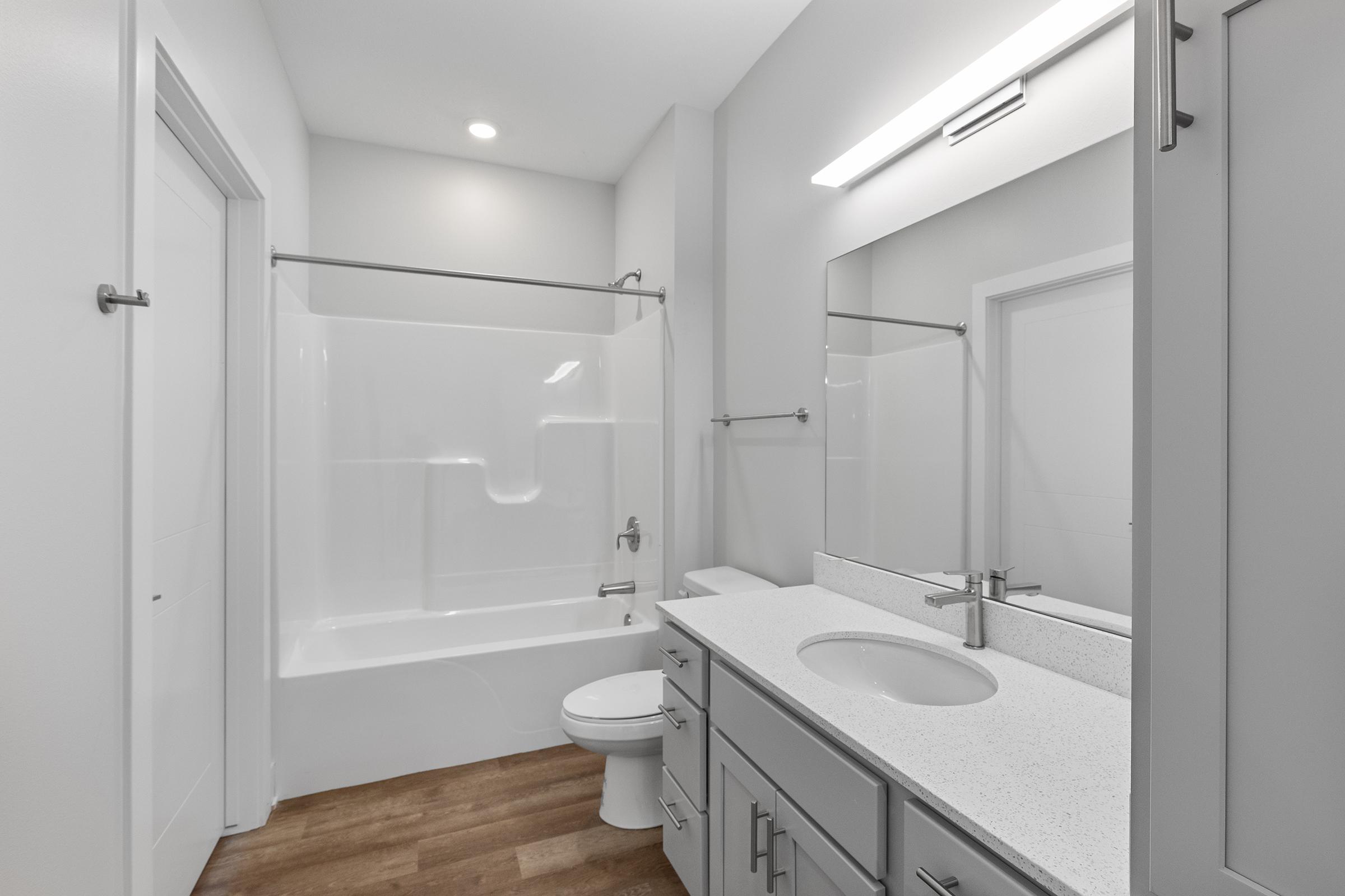 A modern bathroom featuring a white shower-tub combo, a single sink vanity with a light gray cabinet, a large mirror, and wooden laminate flooring. The space is well-lit with overhead lighting, and the walls are painted in a light color, creating a clean and spacious feel.