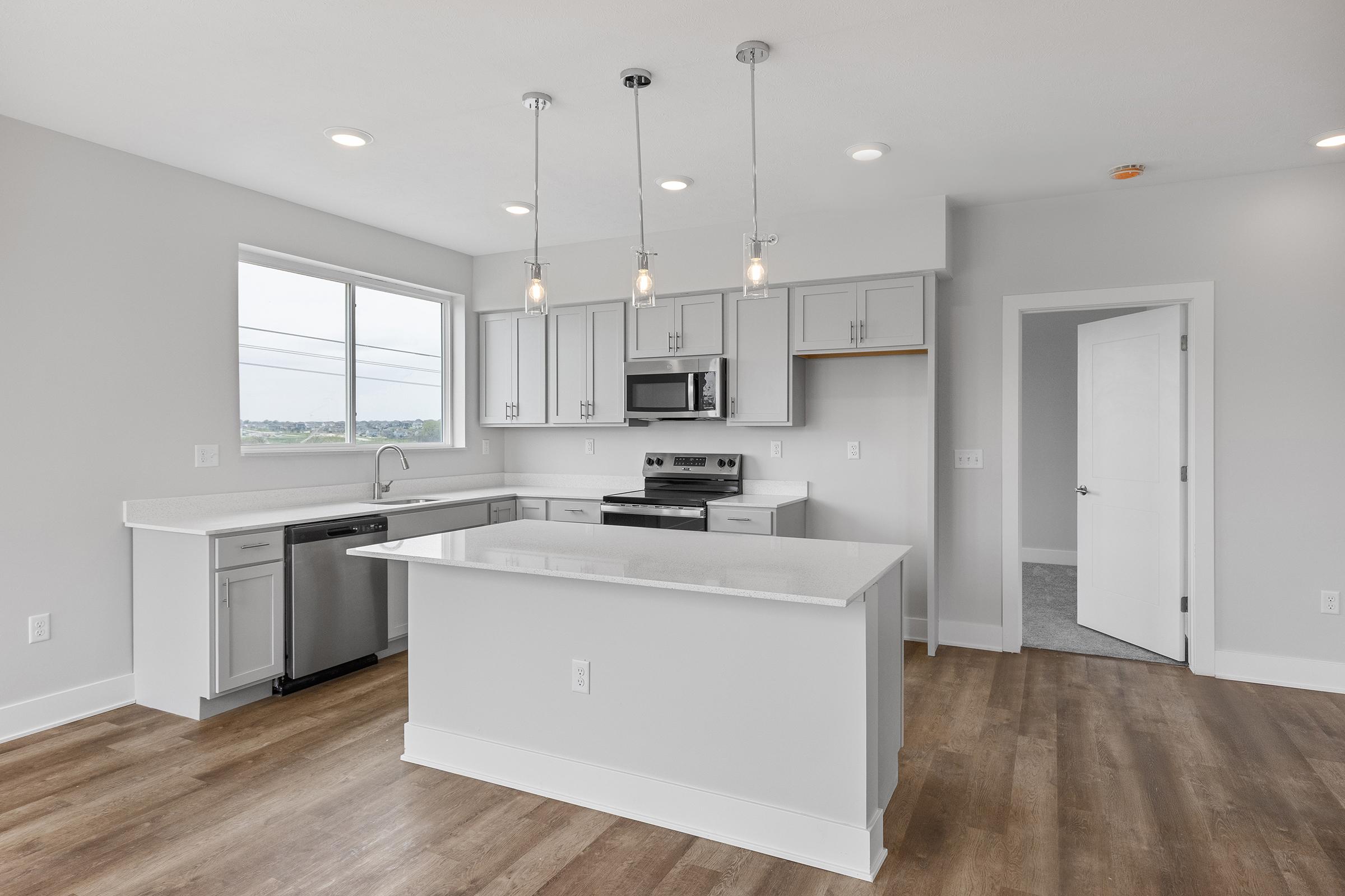 Modern kitchen featuring gray cabinets, stainless steel appliances, and a large island with a white countertop. The area has natural light coming through a window, and stylish pendant lights hang above the island. The flooring is a warm wood tone, adding a cozy atmosphere to the space.