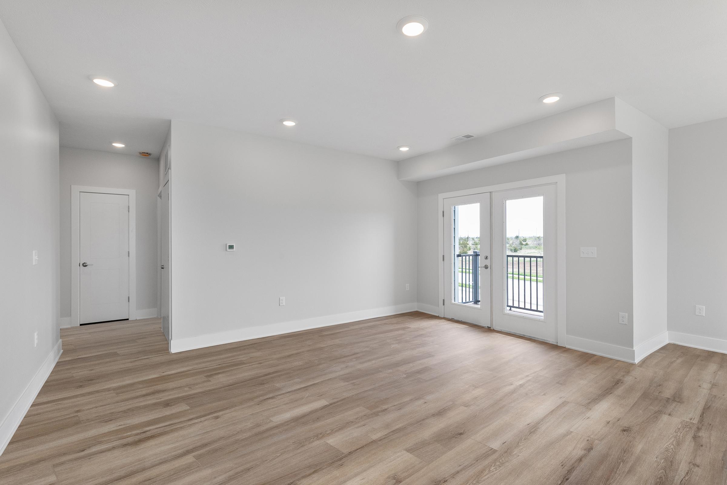 Spacious living area with light-colored walls, hardwood flooring, and large windows. Includes a set of double French doors leading to a balcony. A hallway with a white door is visible on the left. Natural light fills the room, creating a bright and airy atmosphere.