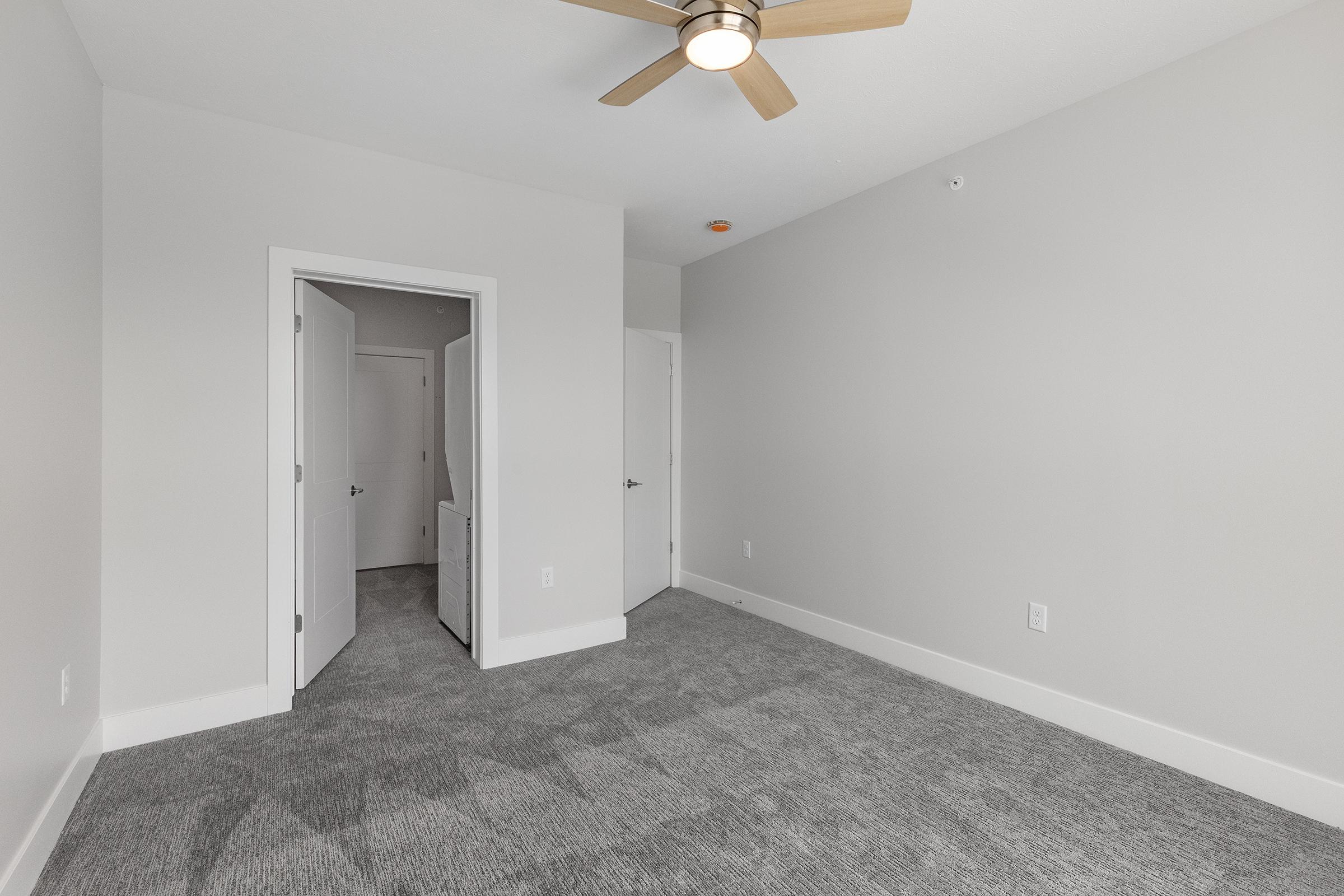 A spacious, light-colored bedroom featuring a ceiling fan, plush gray carpet, and two doorways. One doorway leads to a closet or bathroom, while the other opens to an adjacent room. The walls are painted in a neutral tone, creating a bright and airy atmosphere.