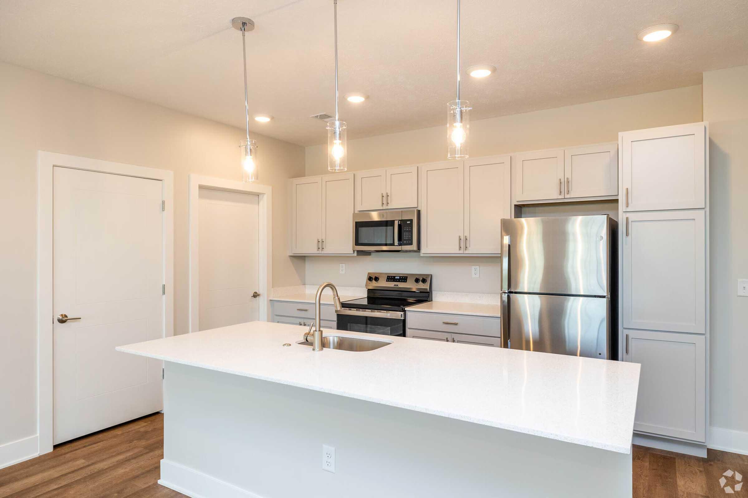 Modern kitchen featuring an island with a sink, light-colored cabinets, stainless steel appliances, and pendant lighting. The space has a clean, contemporary design with wooden flooring and a light, airy feel.