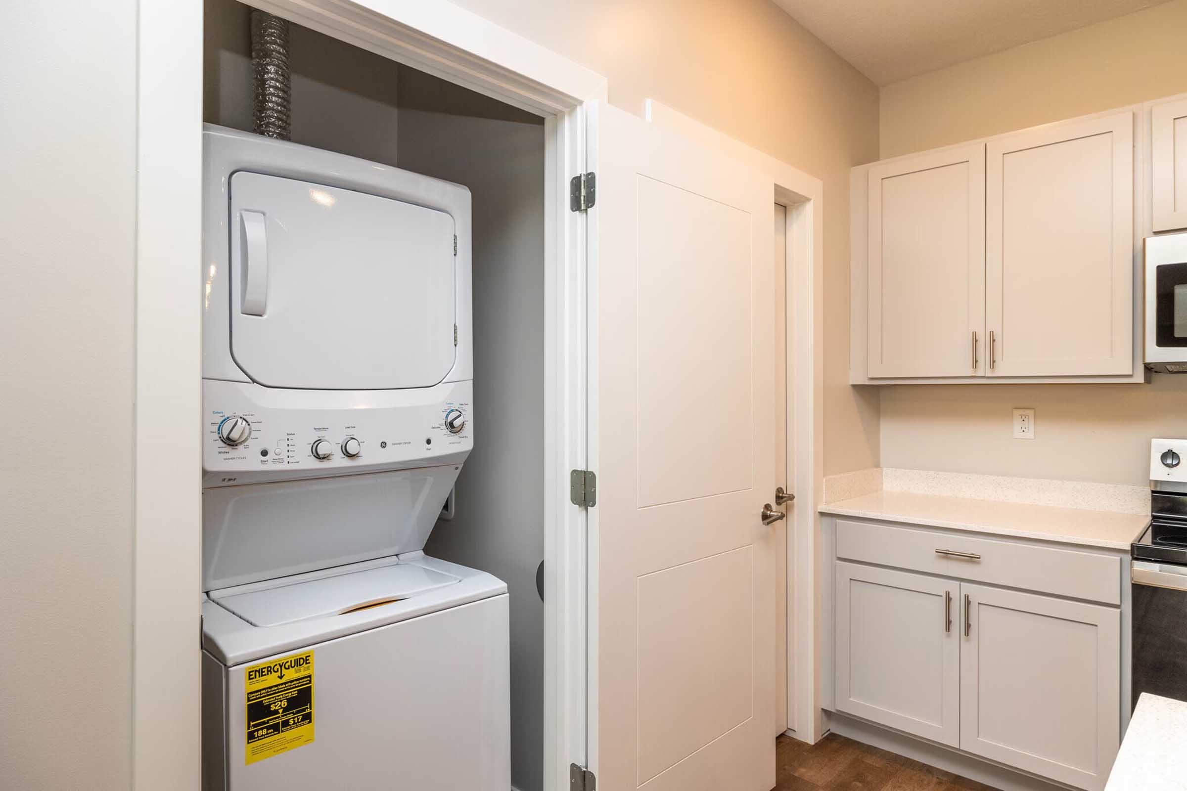 A compact laundry area featuring a stacked washer and dryer. The appliances are located in a small nook near a white door and modern kitchen cabinetry, with a light-colored countertop and silver microwave. The overall design is contemporary and functional.