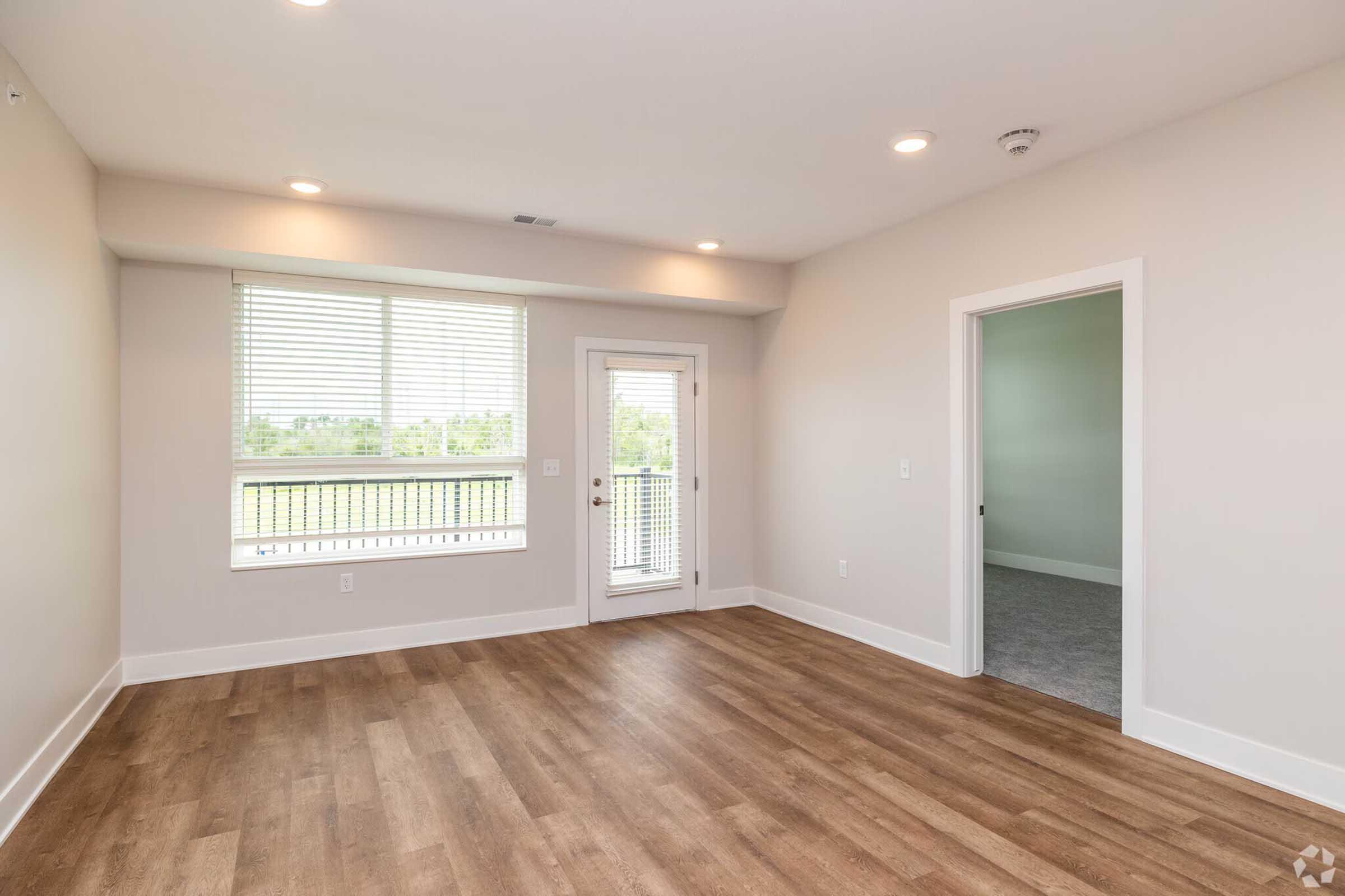 An interior view of a spacious, empty room with light-colored walls and wooden flooring. It features a large window with blinds, a glass door leading to a balcony, and a neutral color scheme. The space is well-lit with overhead lighting and has a clean, modern aesthetic.