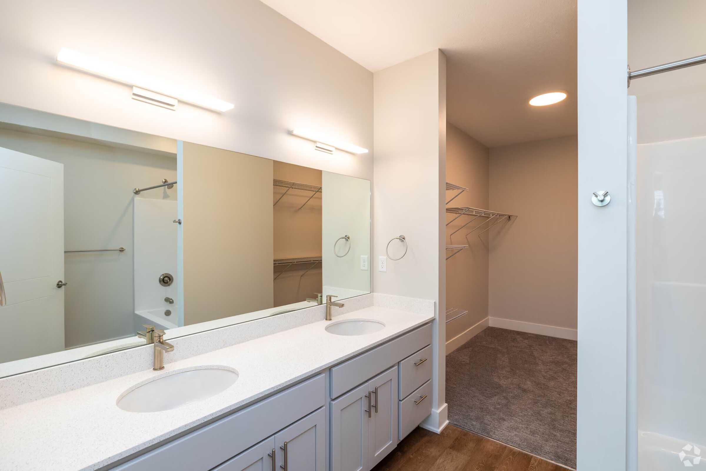 A modern bathroom featuring double sinks with gold faucets, a large wall mirror, and light gray cabinetry. The space includes an adjacent closet area with open shelving and a shower stall. The overall design is clean and contemporary with neutral colors and ample lighting.