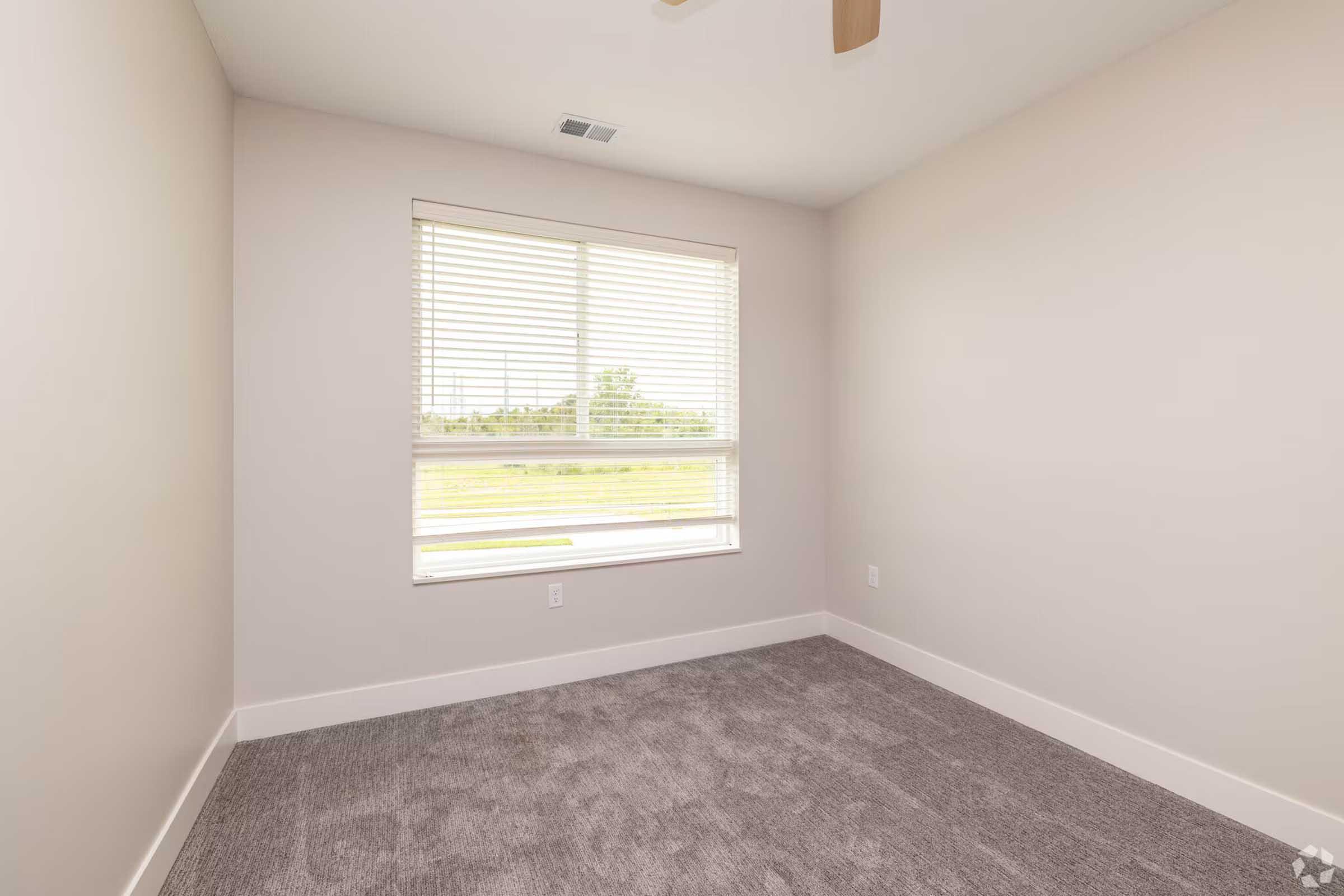 Empty room with light gray carpet, beige walls, and a window featuring white horizontal blinds. Natural light is coming through the window, illuminating the space, and there is a ceiling fan above. The overall atmosphere is bright and inviting.