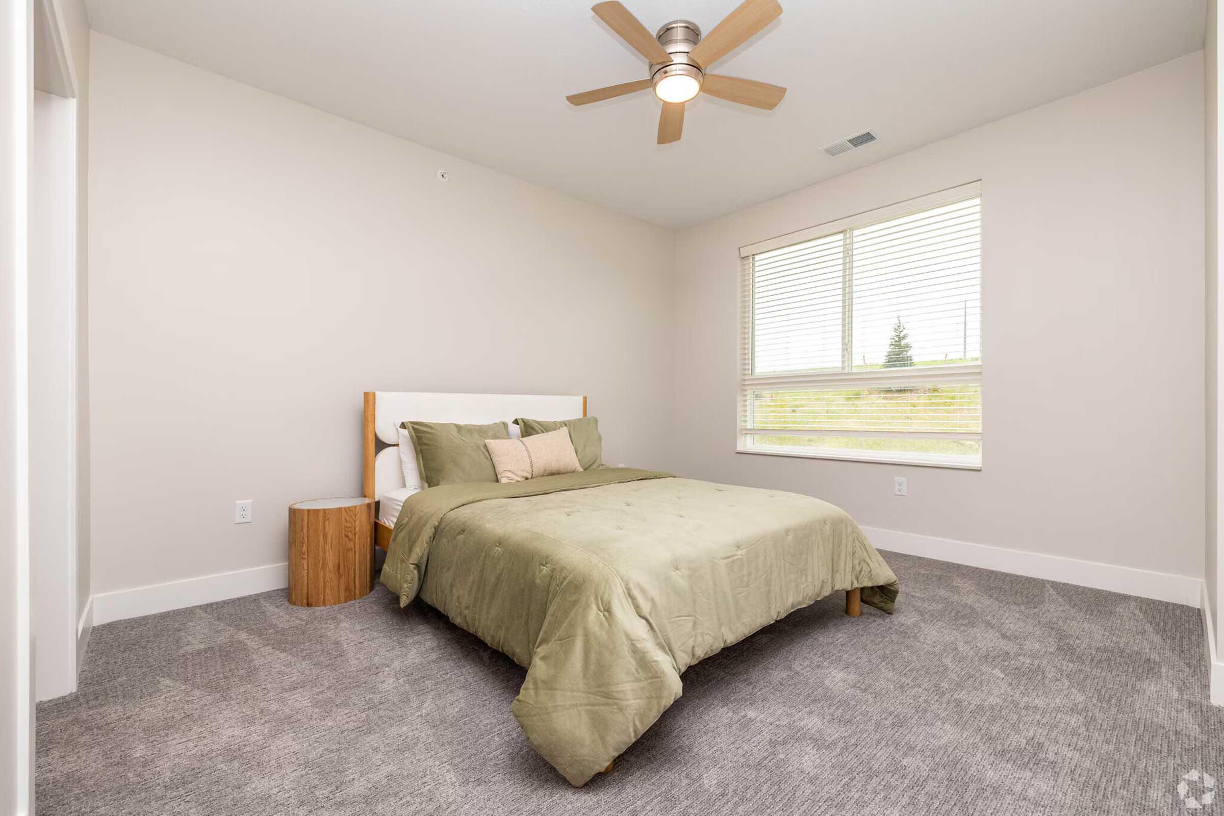 A modern bedroom featuring a queen-sized bed with a green comforter and decorative pillows. There is a wooden nightstand beside the bed, a ceiling fan above, and a window allowing natural light to enter. The walls are light-colored, and the floor is covered with gray carpeting.