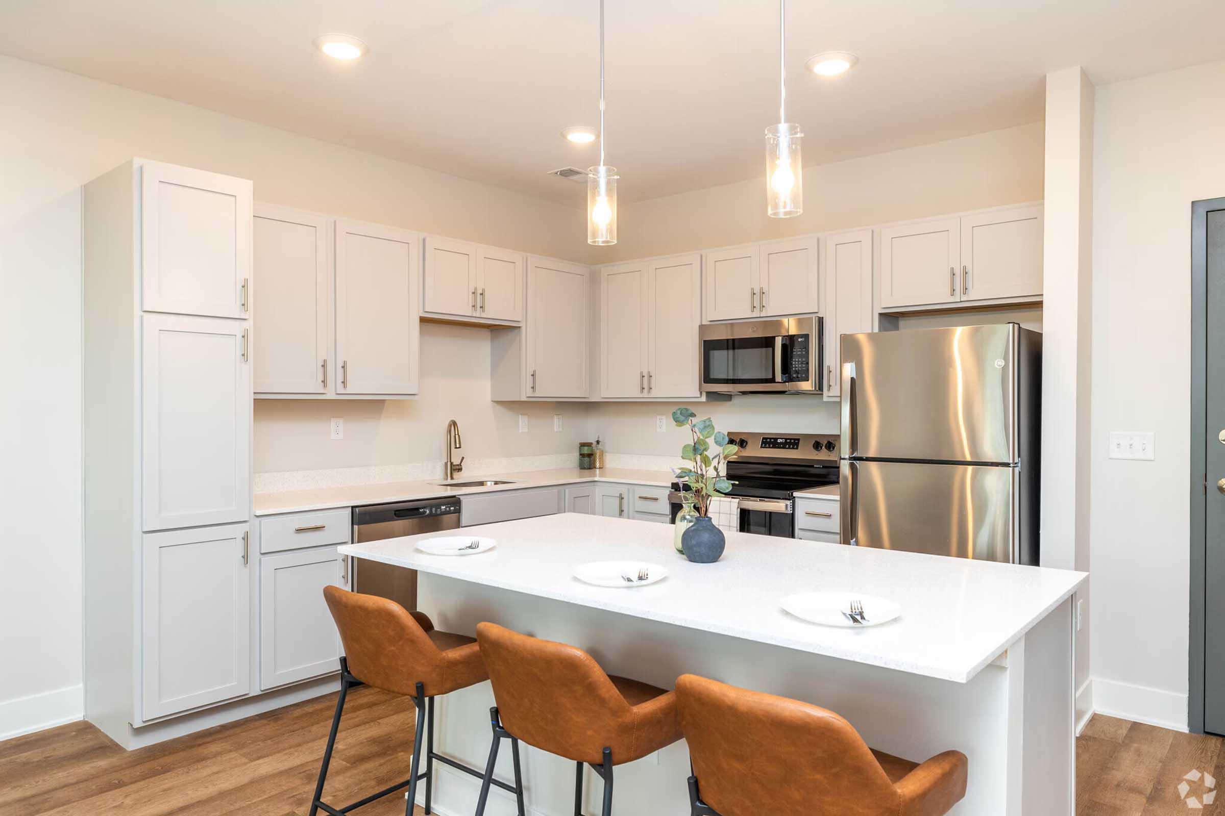 Modern kitchen featuring light gray cabinetry, stainless steel appliances, and quartz countertops. A spacious island with three brown leather bar stools. Pendant lights hang above the island, and a small decorative plant adds a touch of greenery. Wood flooring complements the contemporary design.