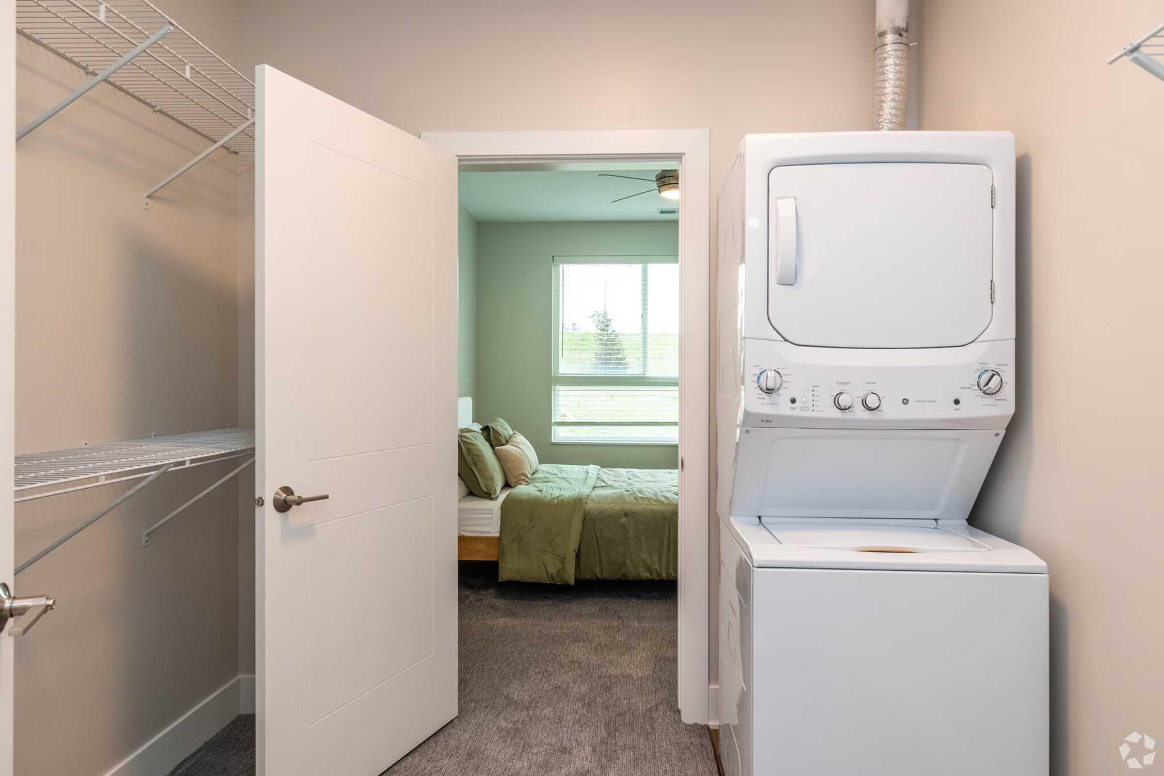 View of a laundry area featuring a stacked washer and dryer, with a door leading into a bedroom in the background. The bedroom includes a bed with green bedding and a window with natural light coming in. The walls are painted a neutral color, and a closet rack is visible on the left side.