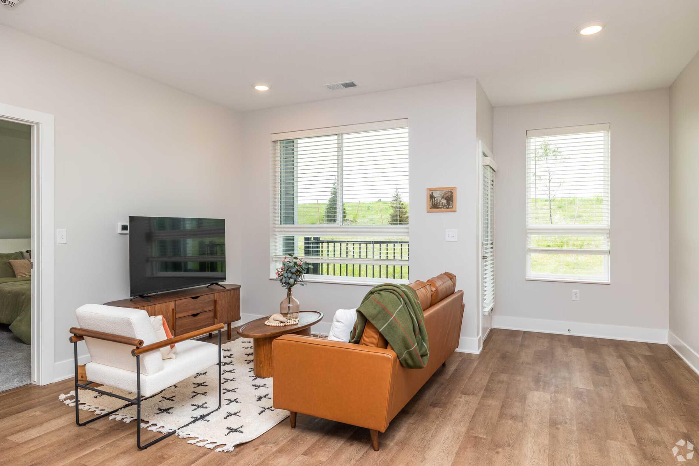 A modern living room featuring a brown leather sofa and a white armchair with a patterned rug. There’s a wooden coffee table with a vase of flowers, a TV on a stand, and large windows allowing natural light. A view of greenery is visible through the windows, creating a bright and inviting atmosphere.