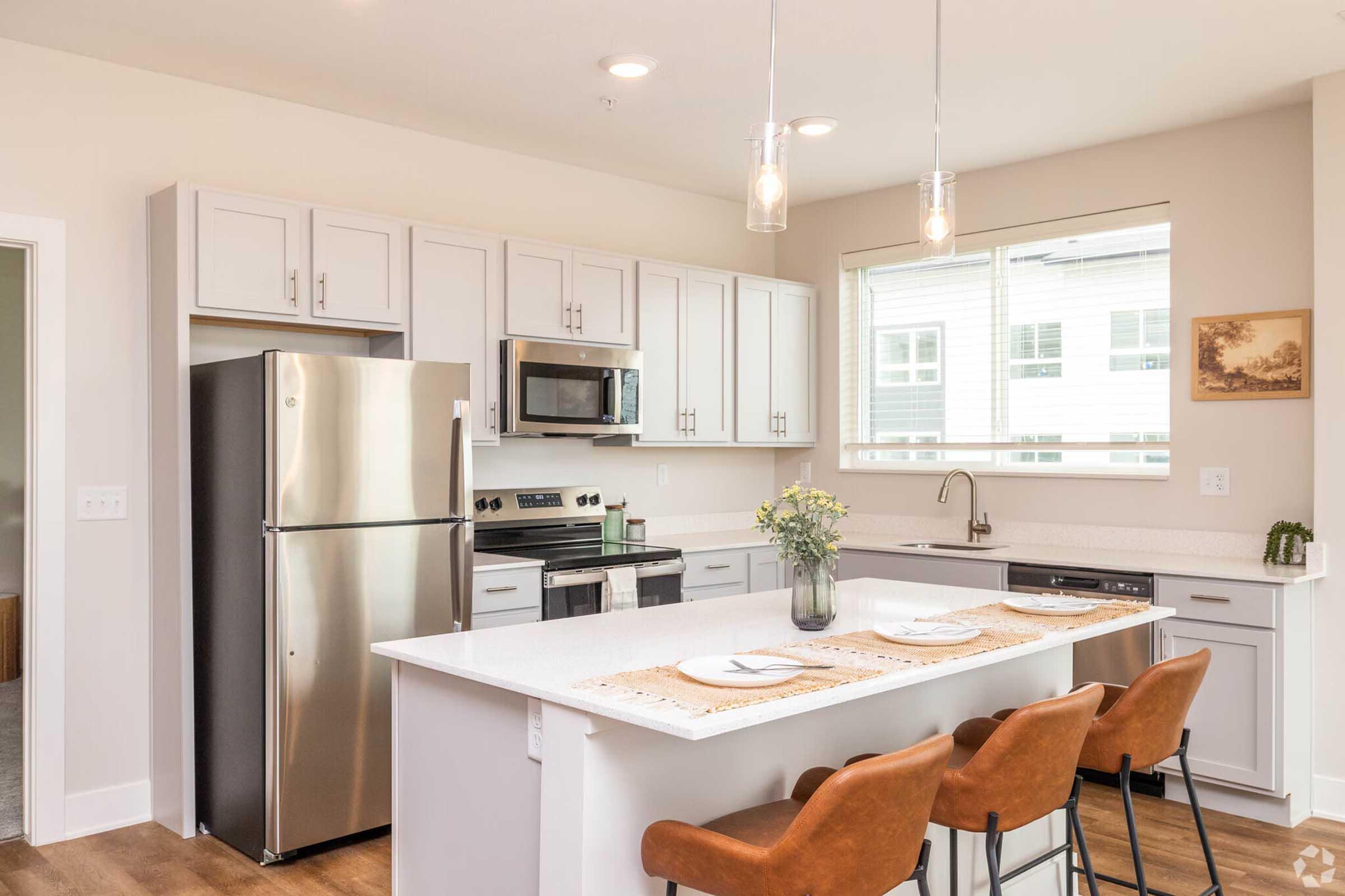 Modern kitchen featuring stainless steel appliances, including a refrigerator and microwave. There is a gray countertop with bar seating for four, and a window providing natural light. The cabinets are light-colored, and the decor includes a small vase of flowers and a framed picture on the wall.