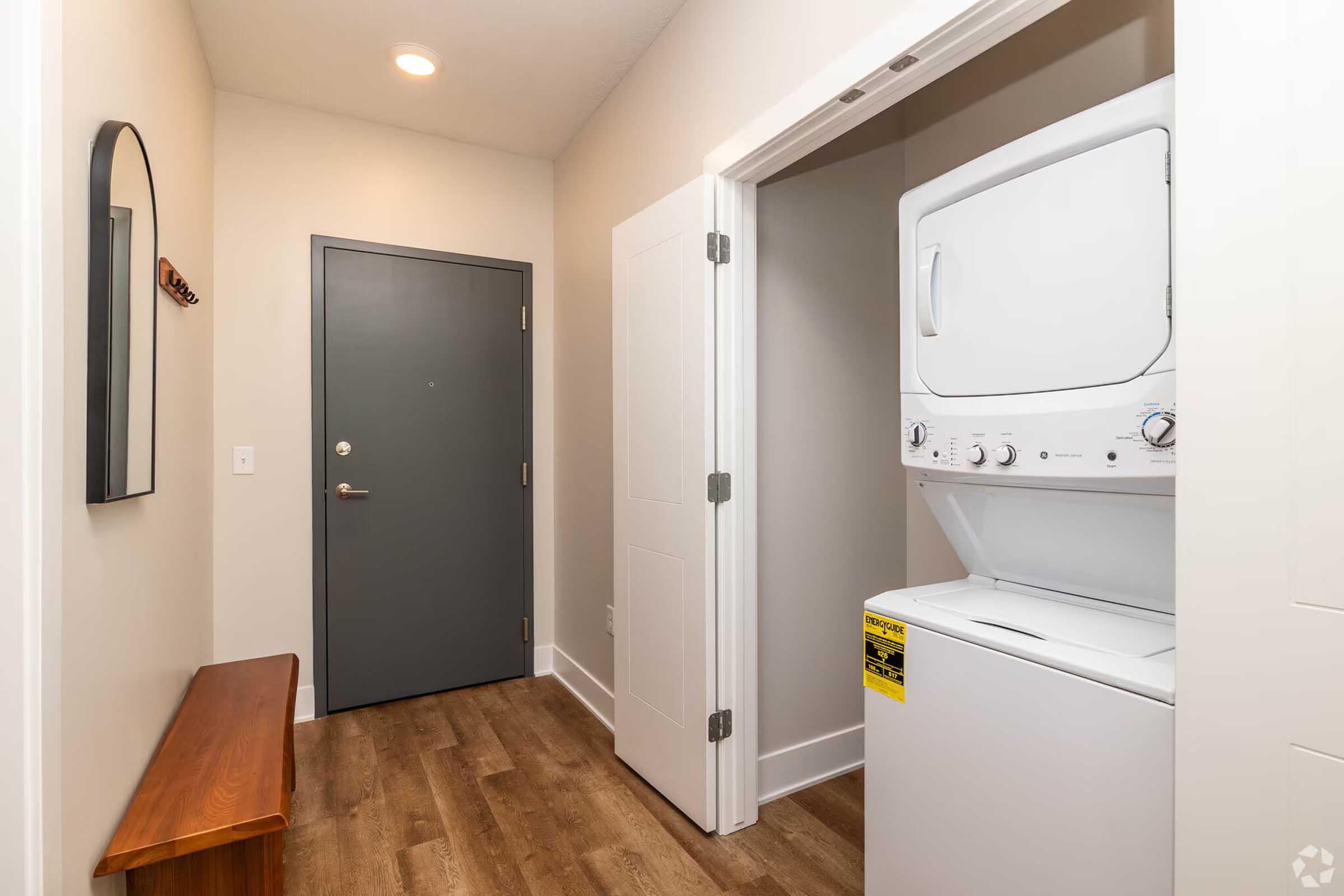 A hallway featuring a stacked washer and dryer on the right, a dark grey door on the left, and a wooden bench against the wall. The floor is made of light-colored wood, and there's a round mirror hanging above the bench. Soft lighting illuminates the space, creating a clean and modern atmosphere.