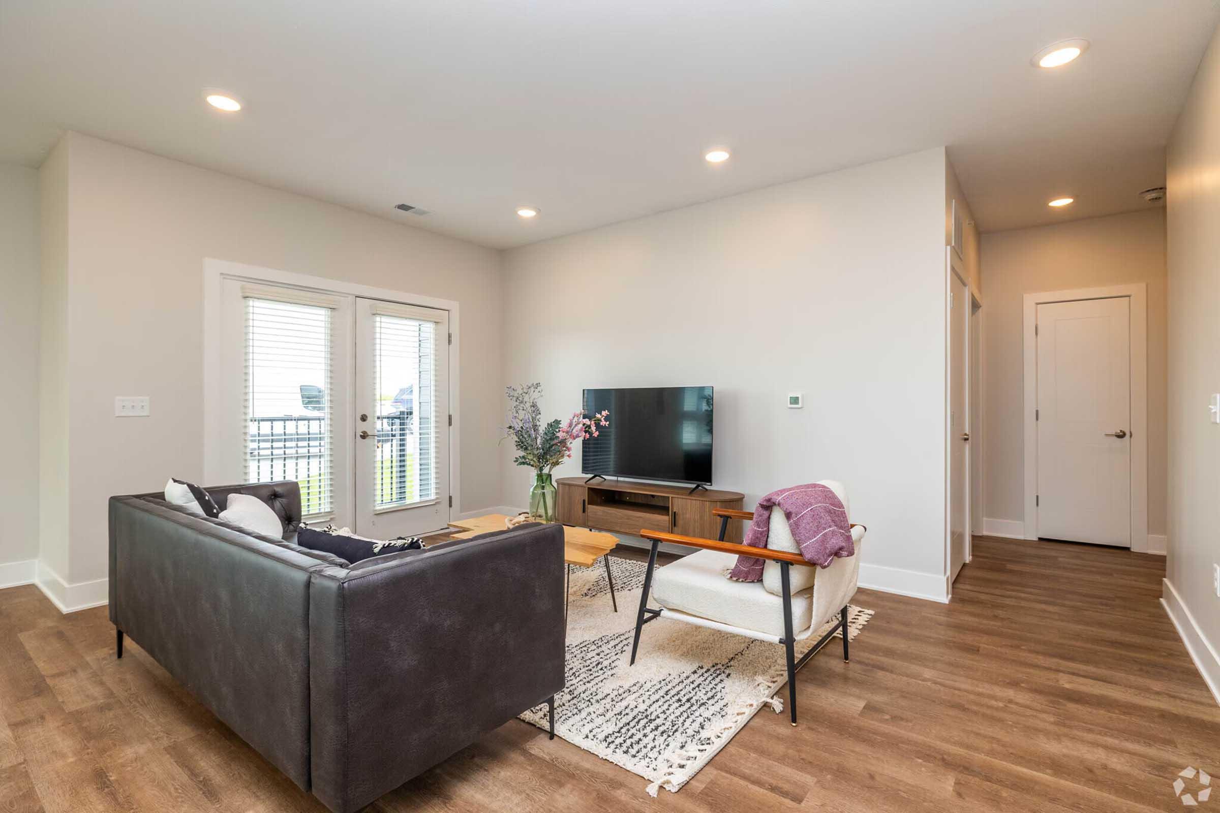 Cozy living room featuring a modern black sofa, a light-colored armchair, a wooden coffee table, and a TV unit. Large windows allow natural light, with access to a balcony. The space has neutral walls and wooden flooring, decorated with a stylish area rug.