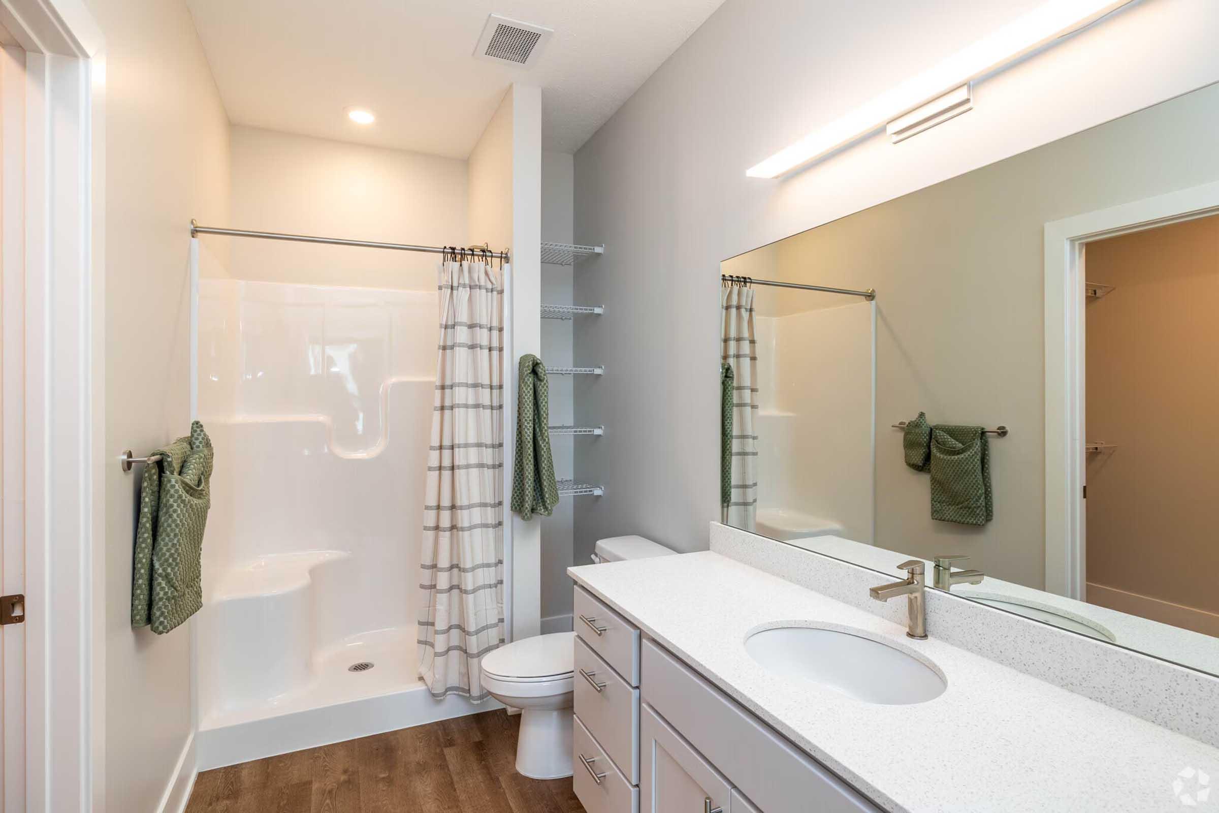 A modern bathroom featuring a white bathtub with a shower curtain, a clean vanity with a sink, a large mirror, and towel racks. The walls are painted in light colors, and there is a wooden floor, adding warmth to the space. Green towels are neatly hung, contributing to the overall fresh and tidy appearance.