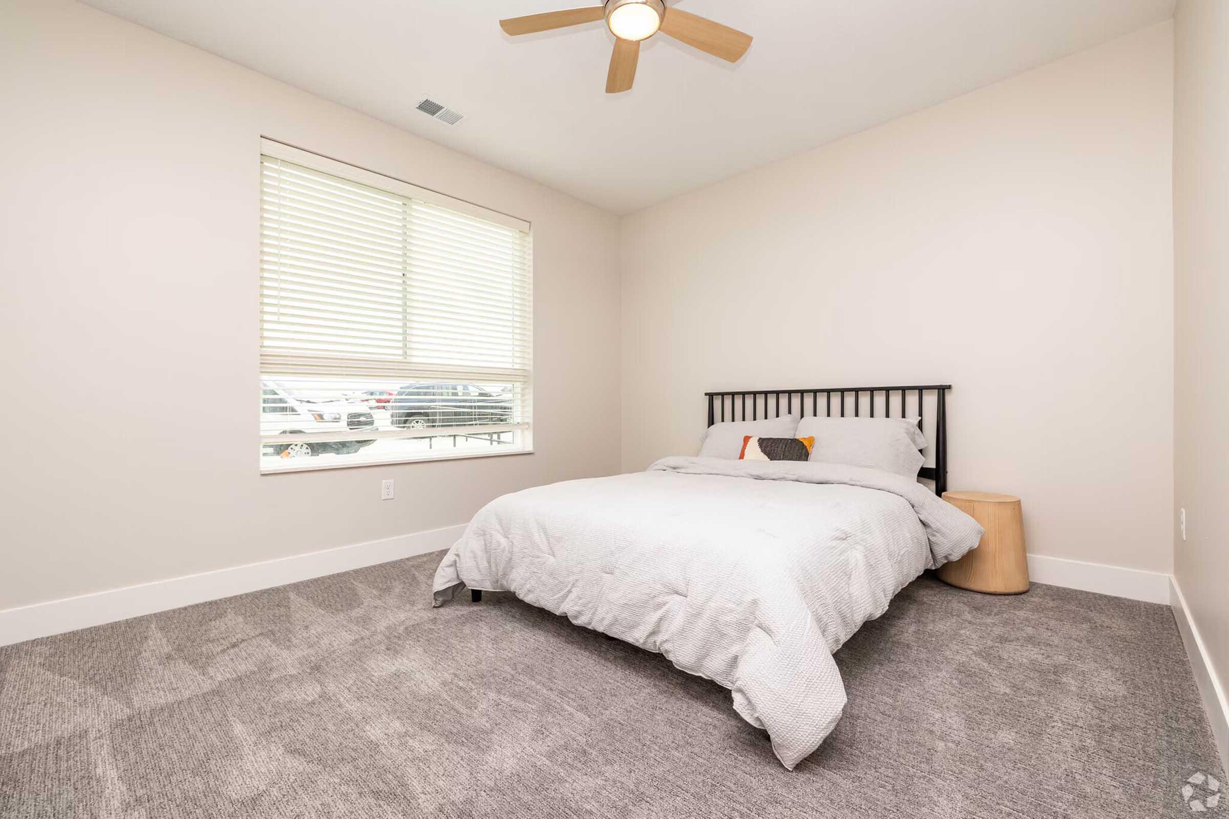 A well-lit bedroom featuring a bed with light gray bedding, a wooden headboard, and a small round side table. The room has a ceiling fan and a window with white blinds, showing a glimpse of the outside. The carpet is light gray, creating a cozy atmosphere.