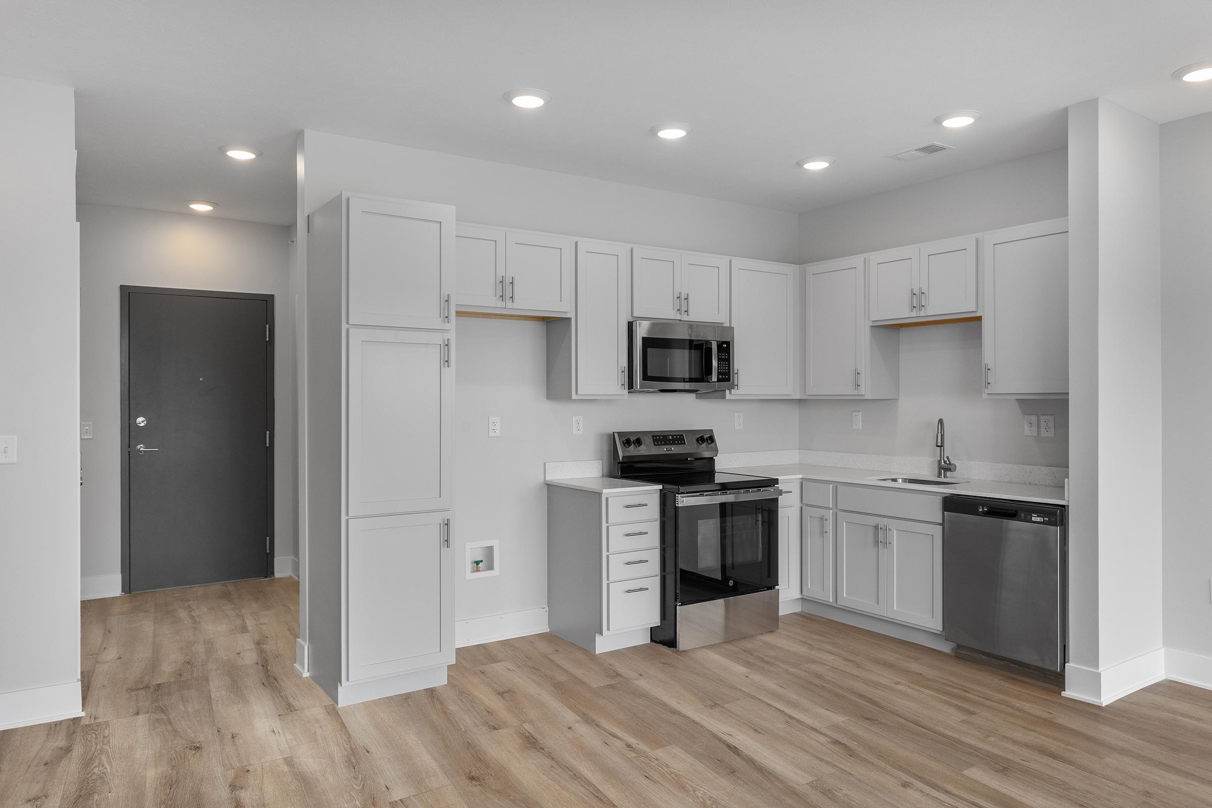 A modern, open-concept kitchen featuring light gray cabinetry, stainless steel appliances including a microwave, oven, and dishwasher, and a white countertop. The floor is light wood, and there are recessed lights in the ceiling. A doorway leads to another room.