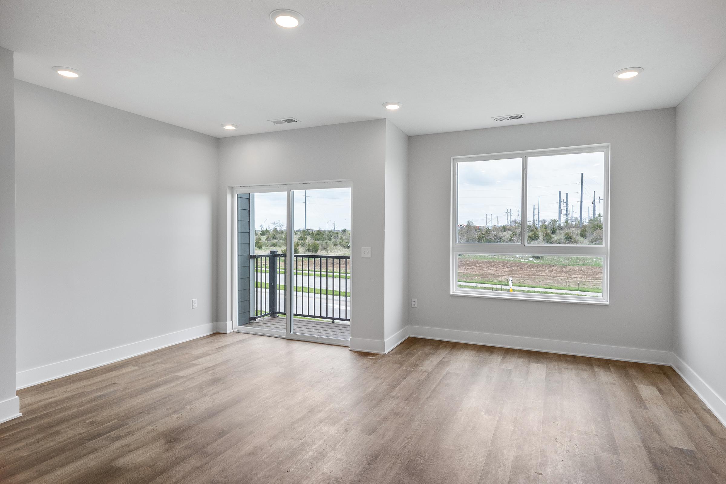 A spacious, empty living room with light gray walls and hardwood flooring. It features large windows allowing natural light, a sliding glass door leading to a balcony, and minimalistic lighting fixtures. The view outside includes greenery and distant power lines.
