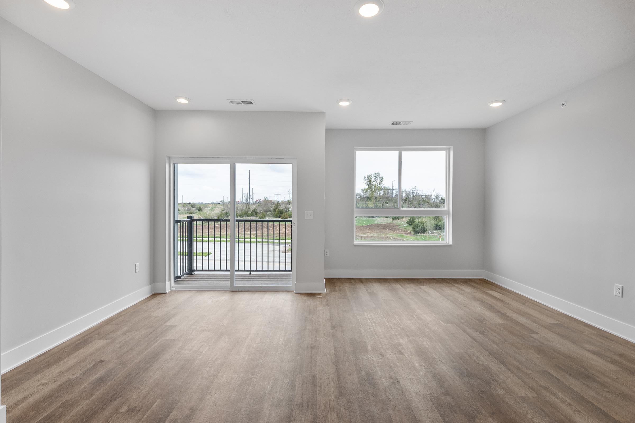 Spacious, empty room with light gray walls and large windows allowing natural light. There is a sliding glass door leading to a balcony with a view of greenery outside. The floor is a light wood finish, and the ceiling has recessed lighting fixtures. Ideal for modern living or office space.