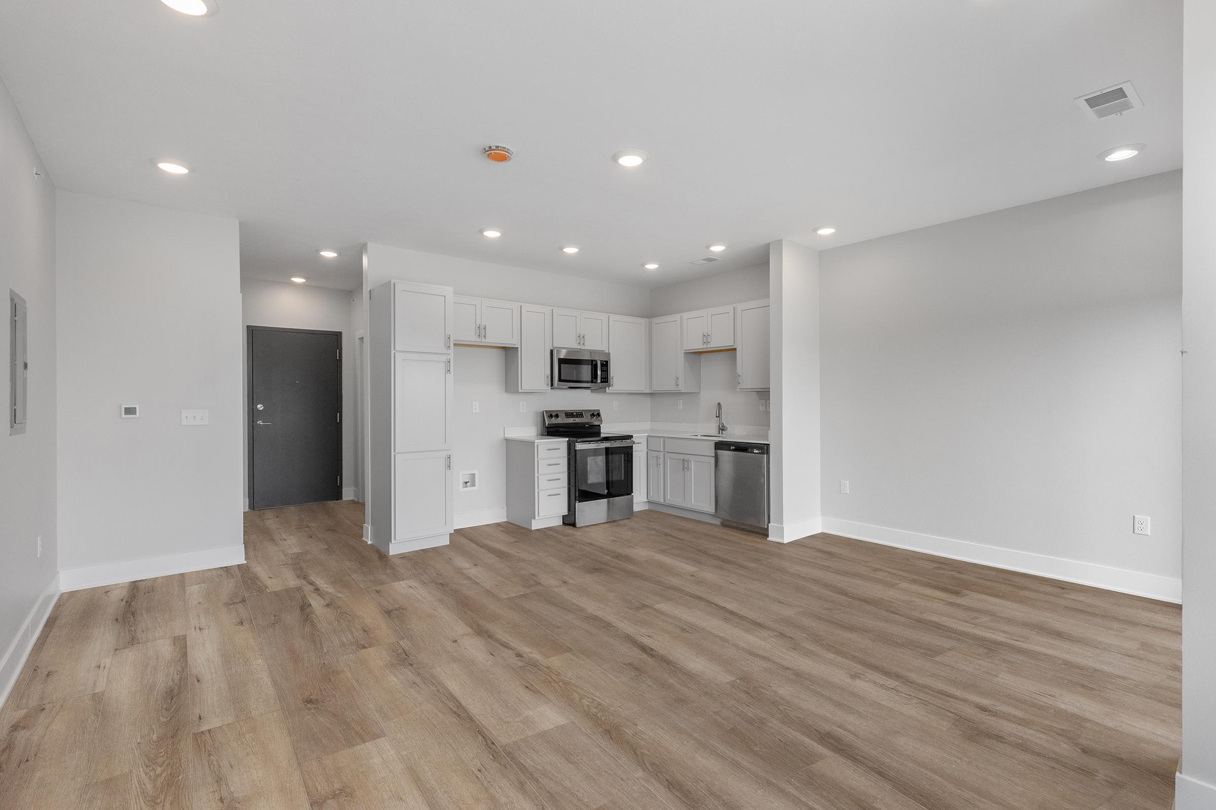 Modern kitchen and living area in an open-plan layout. The kitchen features white cabinets, stainless steel appliances, and a large window allowing natural light to illuminate the space. The floor is light wood, creating a warm atmosphere. The walls are painted in a neutral tone.