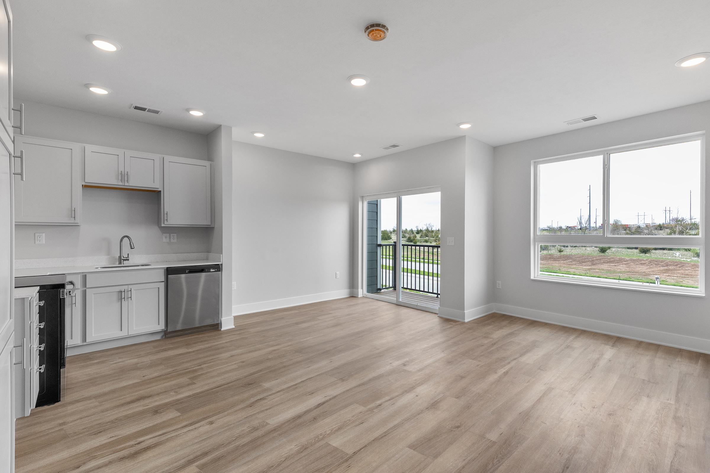 Spacious modern apartment interior with white cabinetry, stainless steel appliances, and large windows offering natural light. The open floor plan features light wood flooring and a sliding glass door leading to a balcony with a view of greenery and industrial structures in the background.