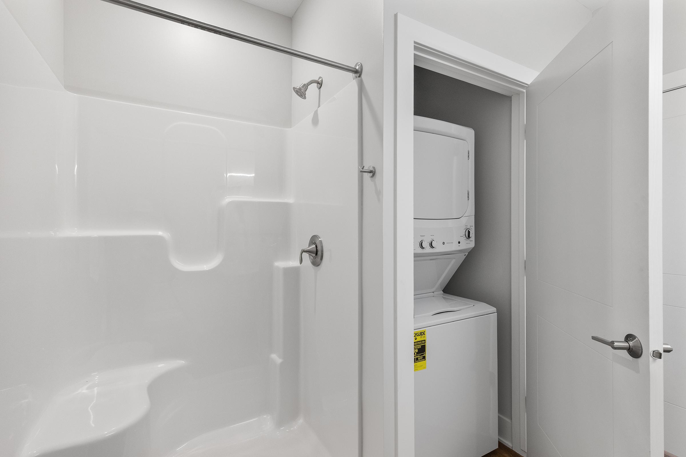 A modern bathroom featuring a clean, white shower stall with a curved design. Adjacent to the shower, a laundry area is visible behind a door, containing a stacked washer and dryer unit. The walls are painted in neutral tones, creating a bright and spacious atmosphere.