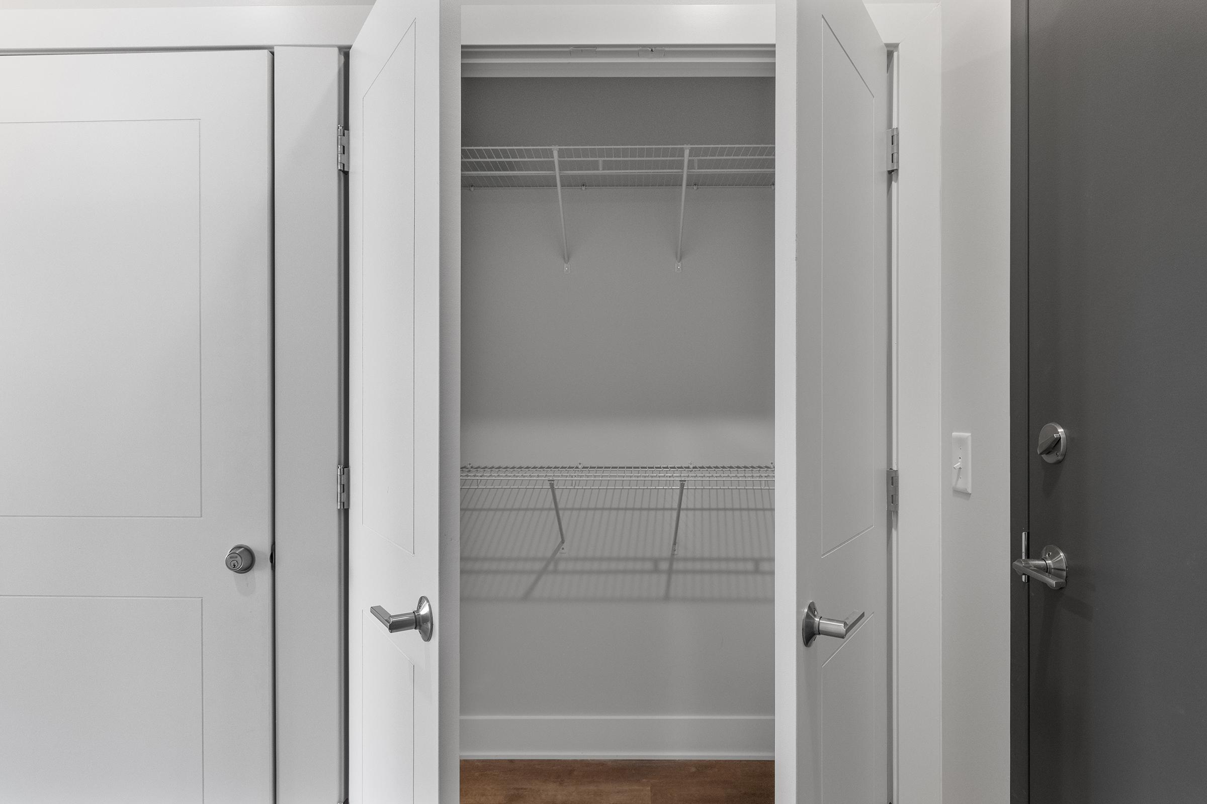 A view of an empty closet with white double doors open, revealing a minimal wire shelving unit inside. The closet has plain walls and a wooden floor, with a door partially visible on the right side.