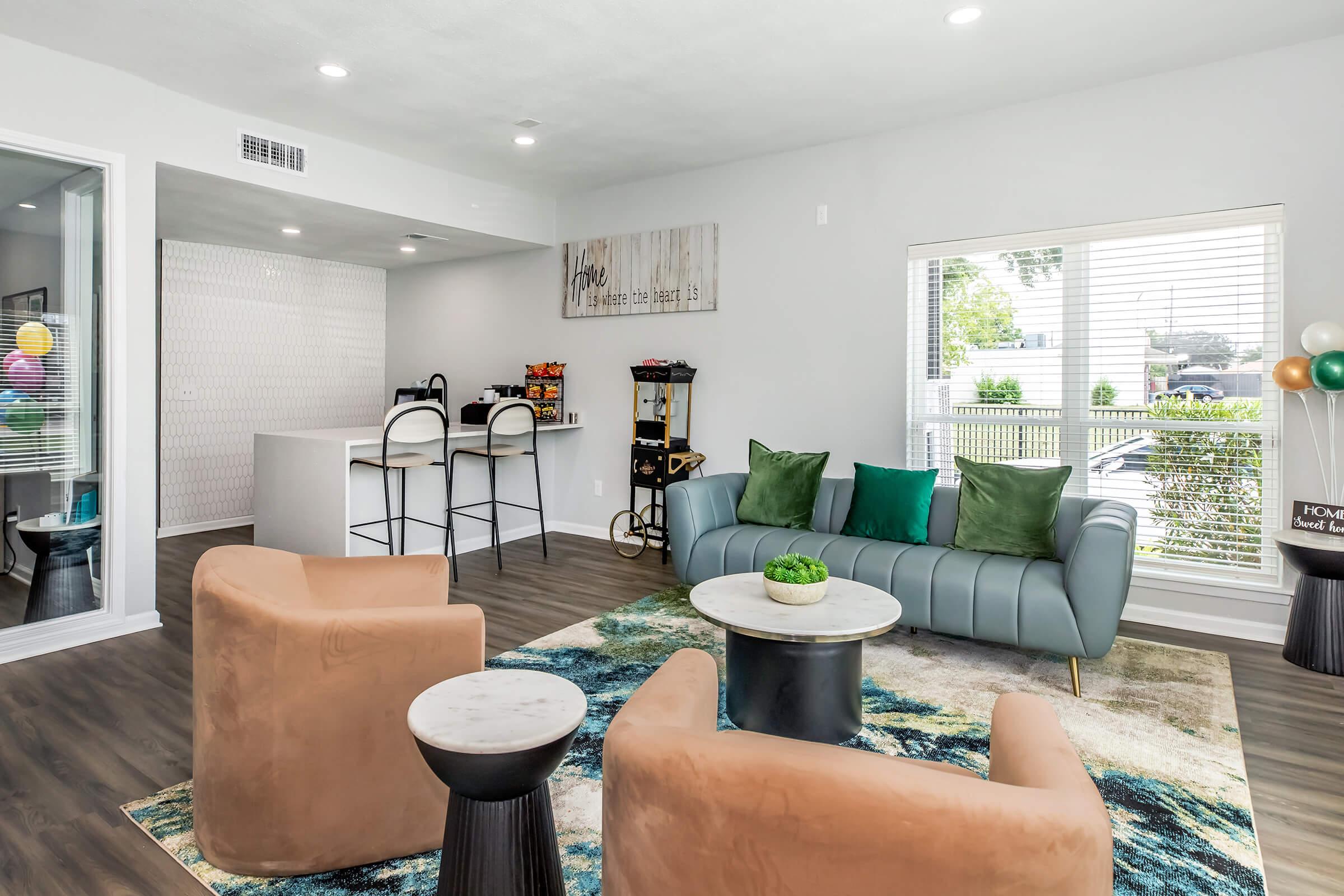 A modern living room featuring two brown accent chairs, a blue sofa with green cushions, and a round coffee table. A patterned rug is beneath the furniture, and there are large windows allowing natural light. In the background, a kitchenette with bar stools is visible along with decorative elements and plants.