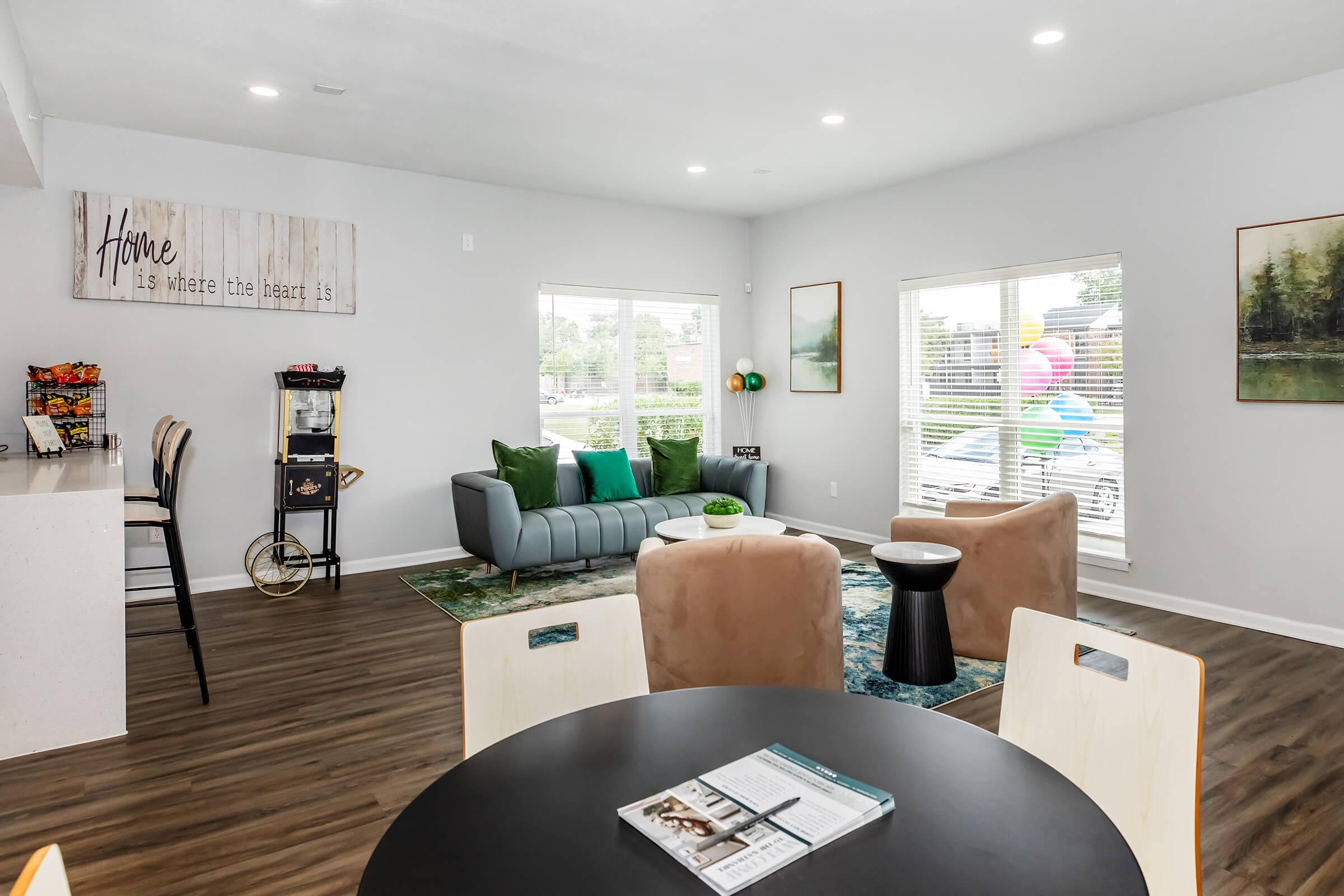 A modern living space featuring a comfortable gray sofa, decorative pillows, and a coffee table. There’s a round black dining table with chairs, and a shelf displaying snacks. Large windows allow natural light, with colorful balloons visible outside. Wall art includes a quote about home and a scenic painting.