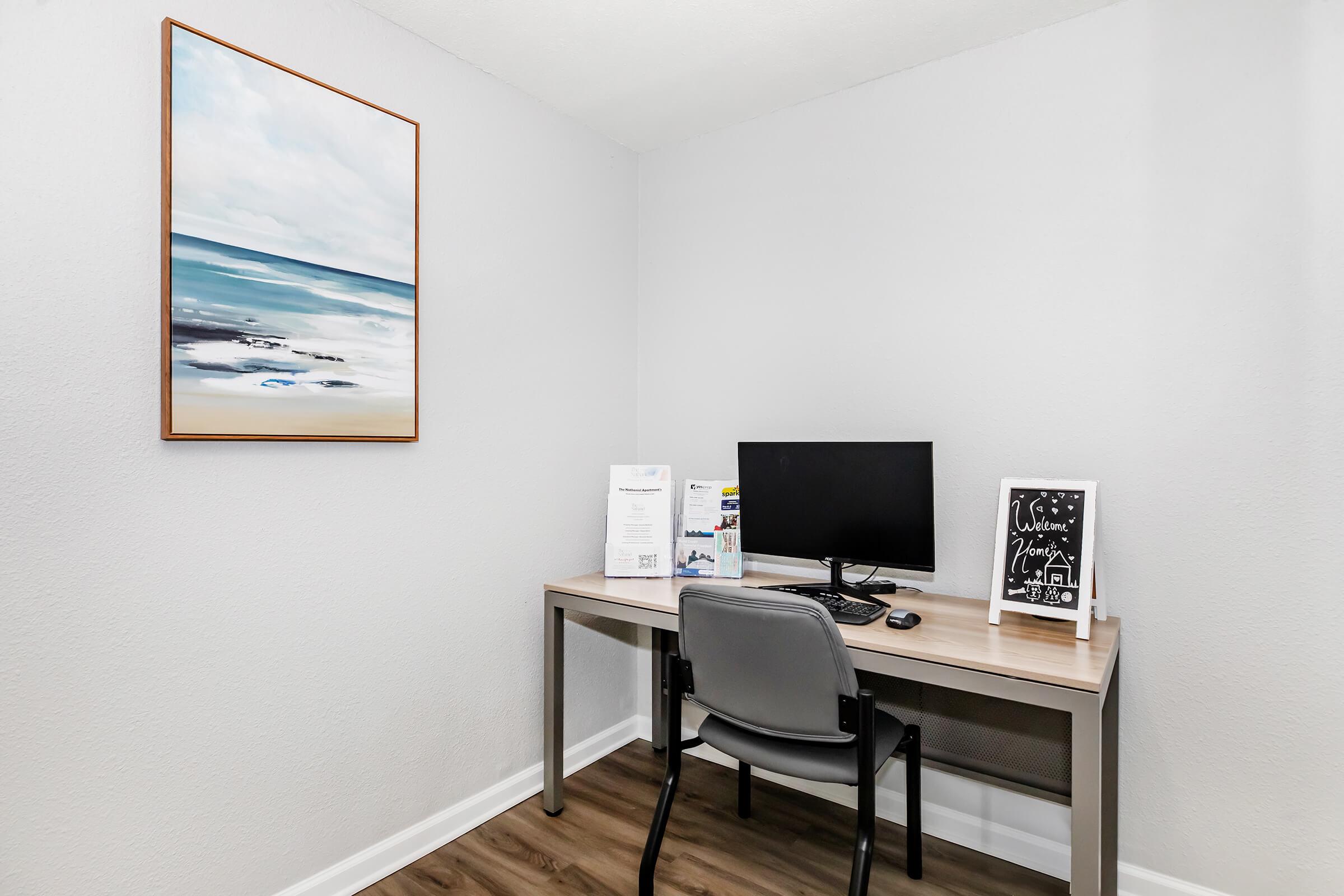A minimalist workspace featuring a wooden desk with a computer monitor, a black chair, and decorative items. There is a framed painting of a seaside scene on the wall, and the room has light gray walls and a wooden floor. The space is organized and bright, designed for productivity.