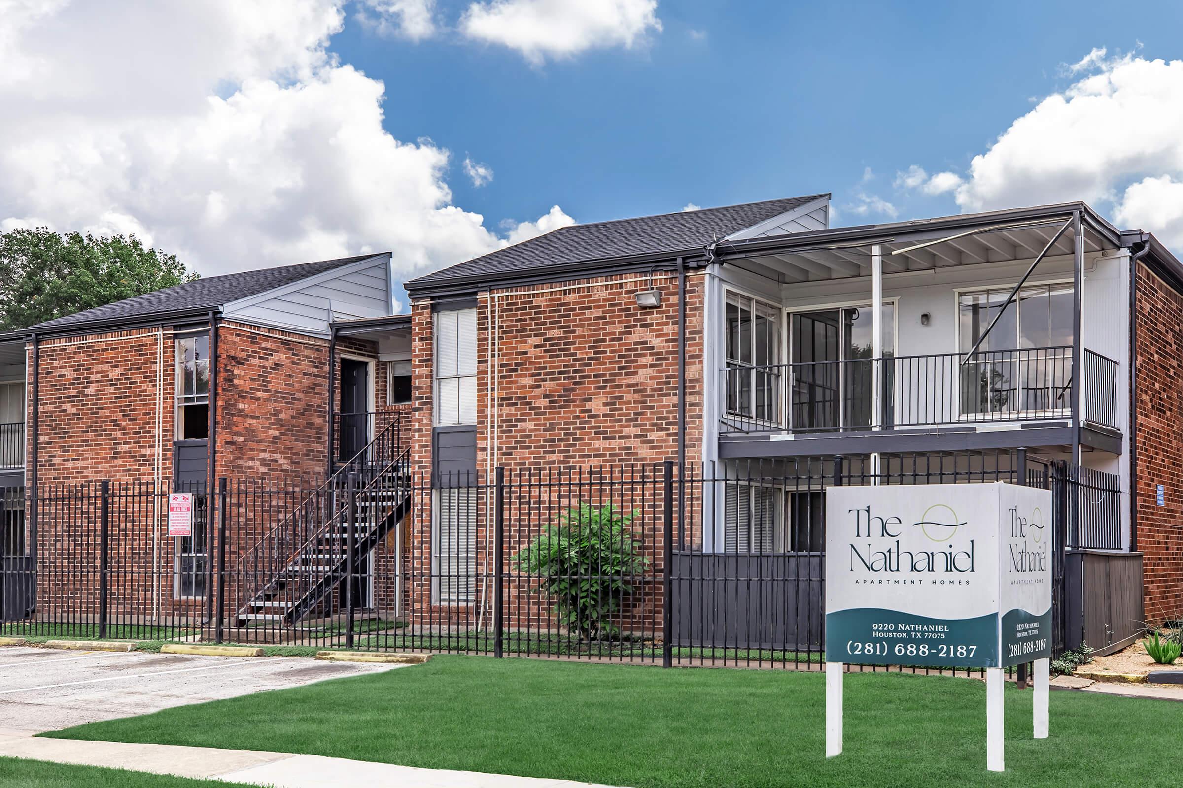 Exterior view of a residential apartment complex named "The Nathaniel." The building features red brick walls, a black roof, and balconies. A sign in front of the building displays contact information. The setting includes green grass and a partly cloudy sky in the background.