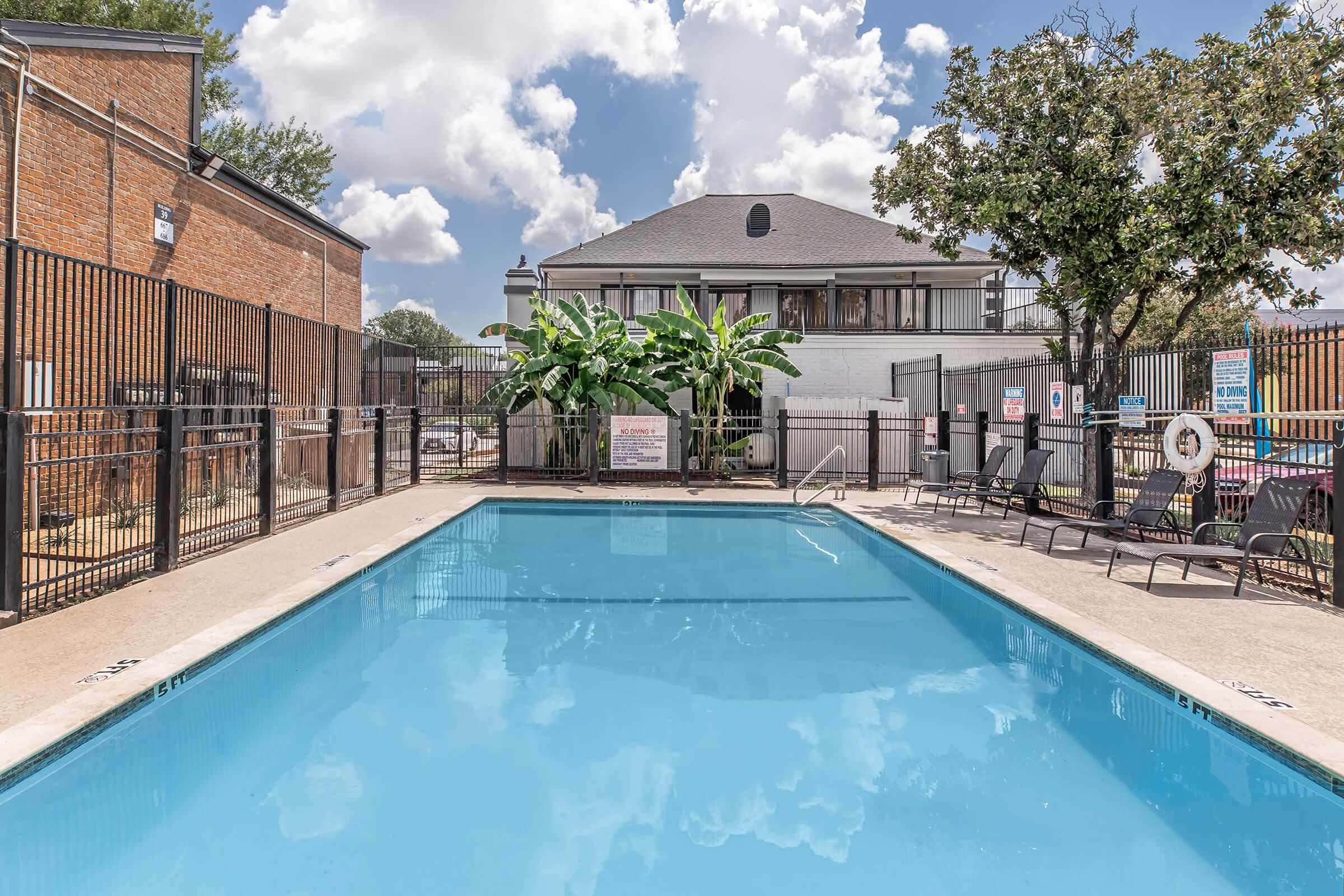 A clean, blue swimming pool surrounded by a black metal fence, with lounge chairs along the edges. In the background, there is a multi-story building and tall plants. The sky is bright with scattered clouds. Signs and safety equipment are visible around the pool area.