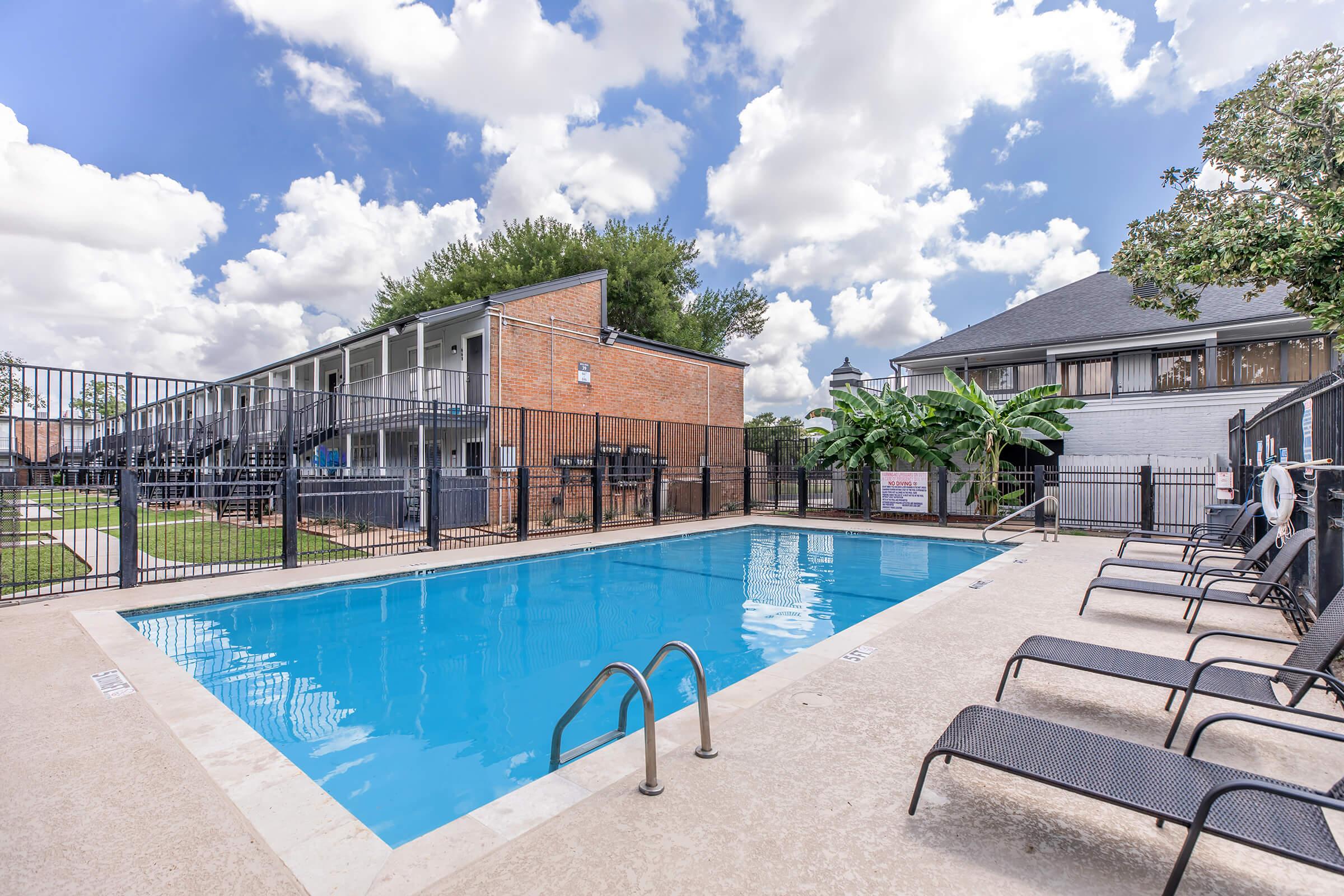 A swimming pool surrounded by lounge chairs at an apartment complex. In the background, there are two buildings, with green trees and a cloudy sky above. The pool area is enclosed with a black fence, creating a peaceful outdoor space.
