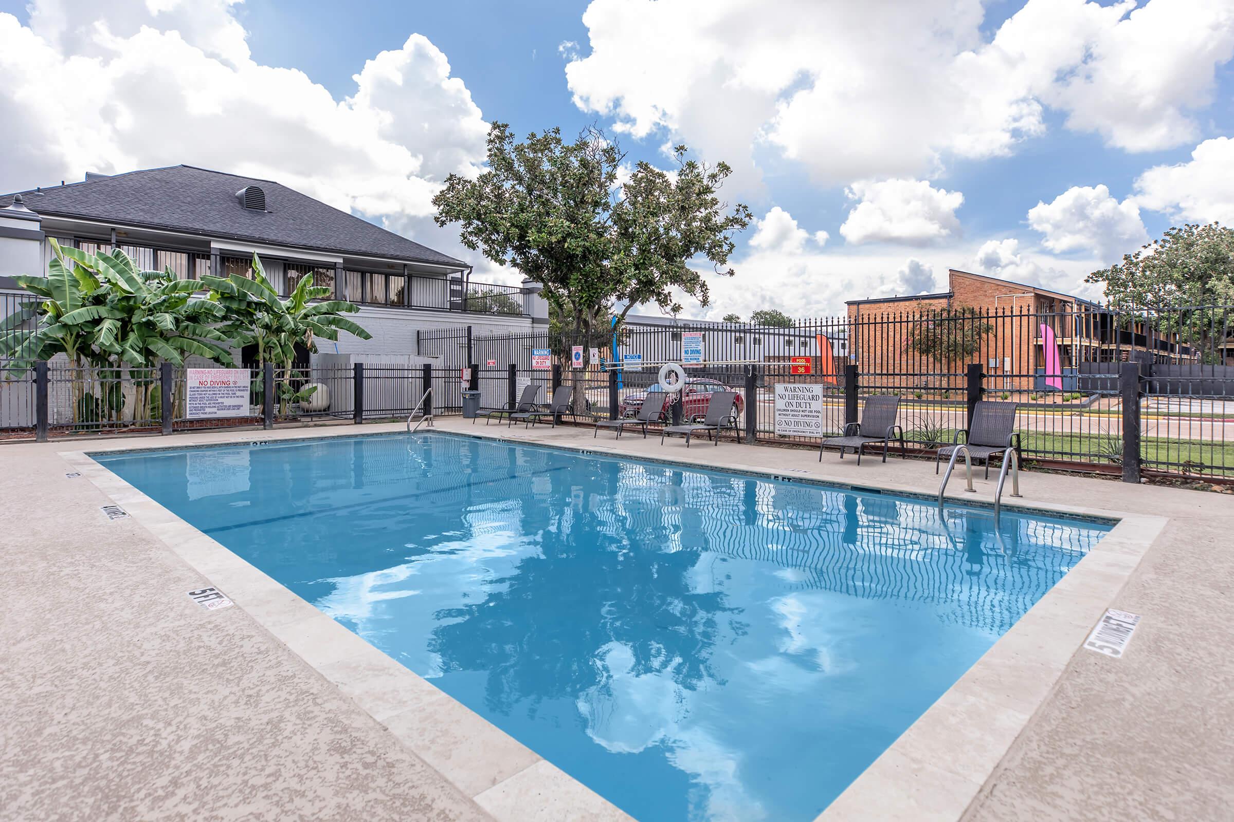 A clear swimming pool surrounded by a fence, with chairs on the side. Lush green plants and trees are visible nearby. The sky is partly cloudy, and there are signs around the pool area. A building is seen in the background, indicating a residential setting.