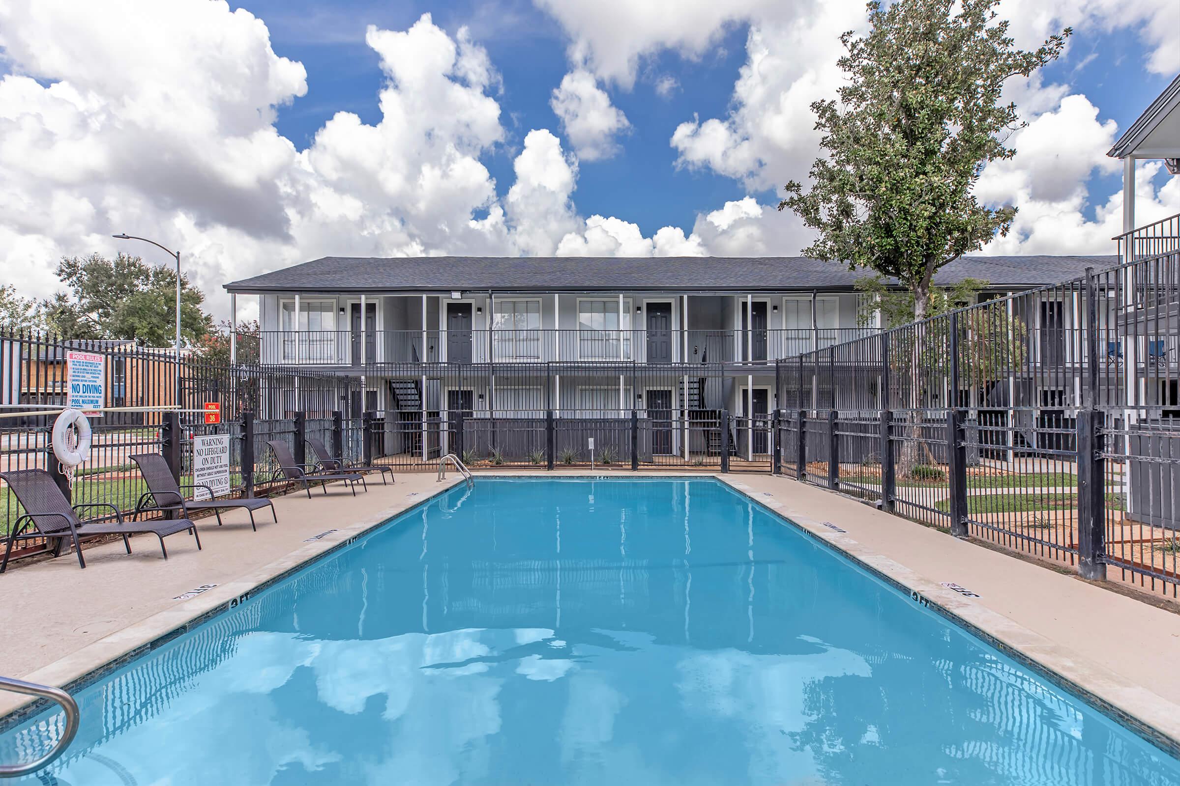 A clear blue swimming pool surrounded by lounge chairs, with a two-story building in the background. The building features multiple balconies and is set against a partly cloudy sky. A tree is visible near the pool area, creating a tranquil and inviting atmosphere.