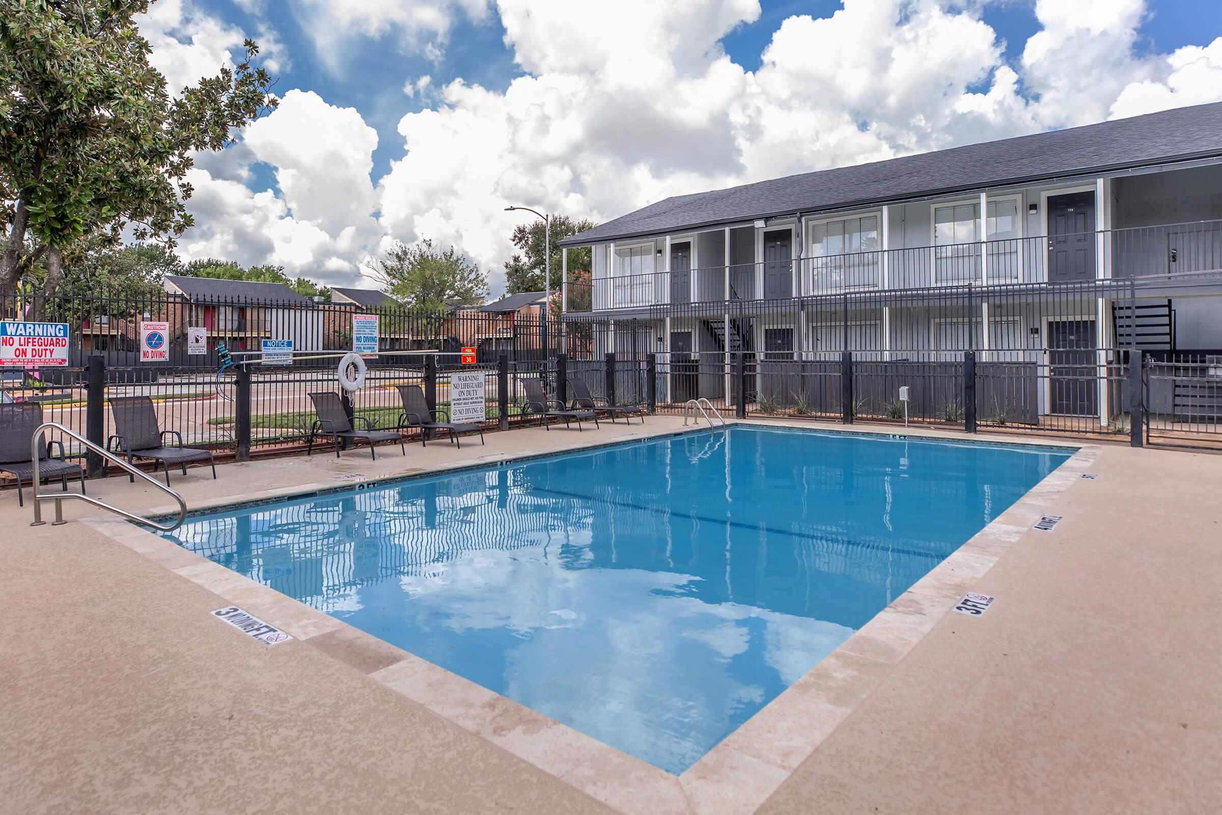 View of a swimming pool surrounded by lounge chairs, with two-story buildings in the background. The pool area is enclosed with a fence, and there are clouds in a partially blue sky. Signs indicating pool rules are visible around the area.