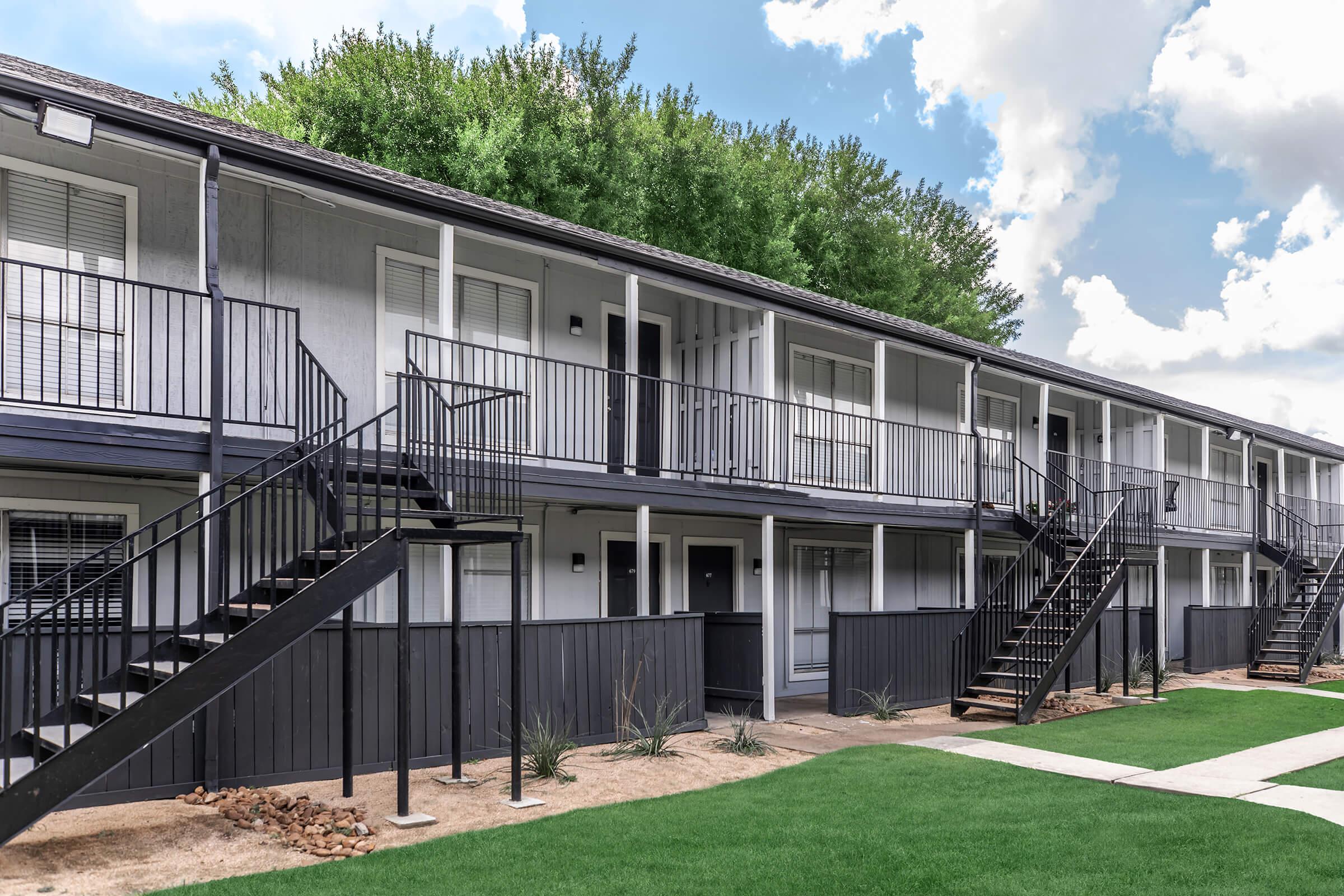 Two-story apartment building featuring multiple units with black railings and stairs. The facade is light-colored with large windows and a grassy area in the foreground. Lush greenery is visible on the roofline, and the sky is partly cloudy.