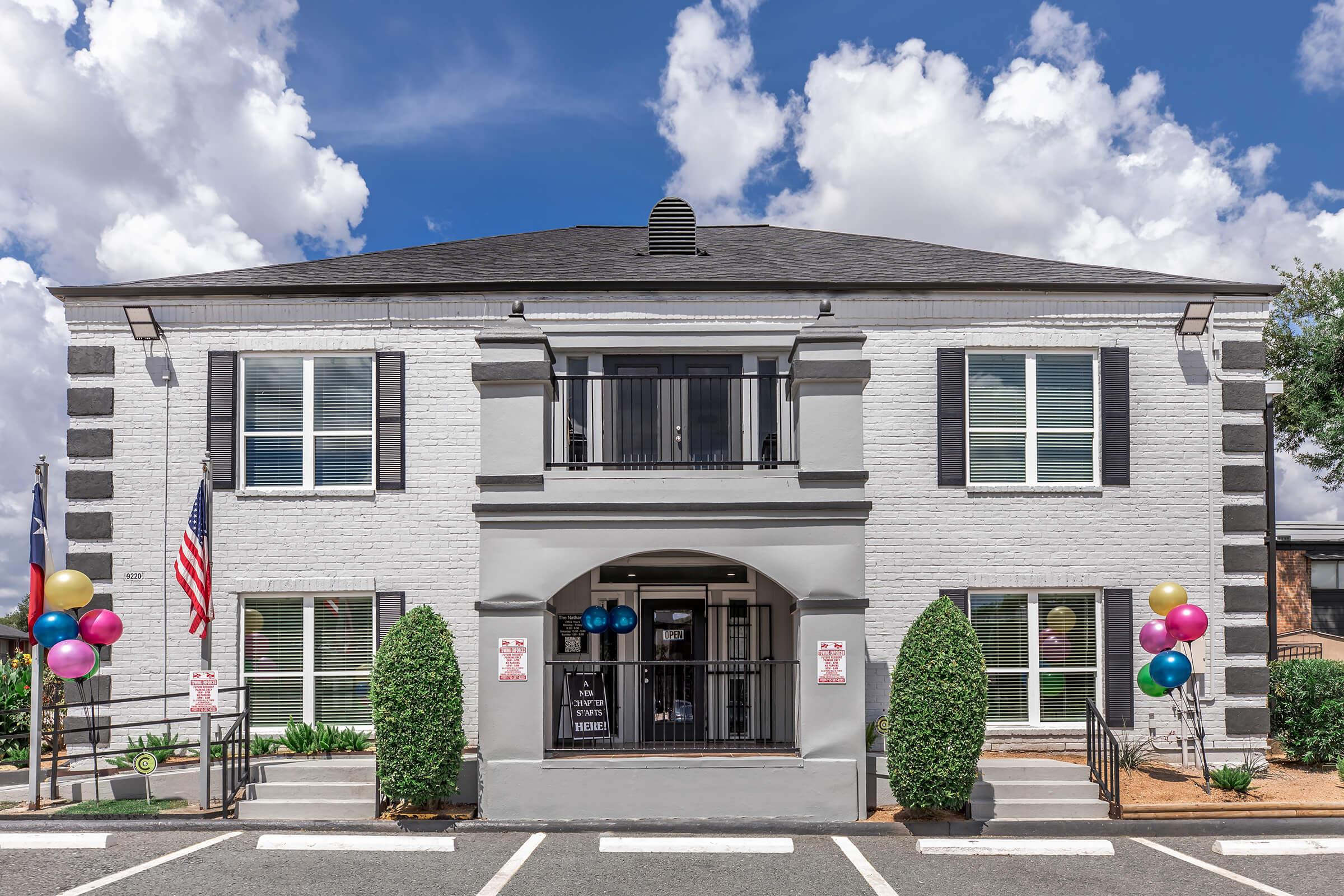 Two-story building with a gray and white exterior, featuring large front windows and a central entrance. Flanking the entrance are two neatly trimmed bushes. Colorful balloons decorate the entrance area, and there are American flags on either side. The sky is partly cloudy. Parking spaces are visible in front.