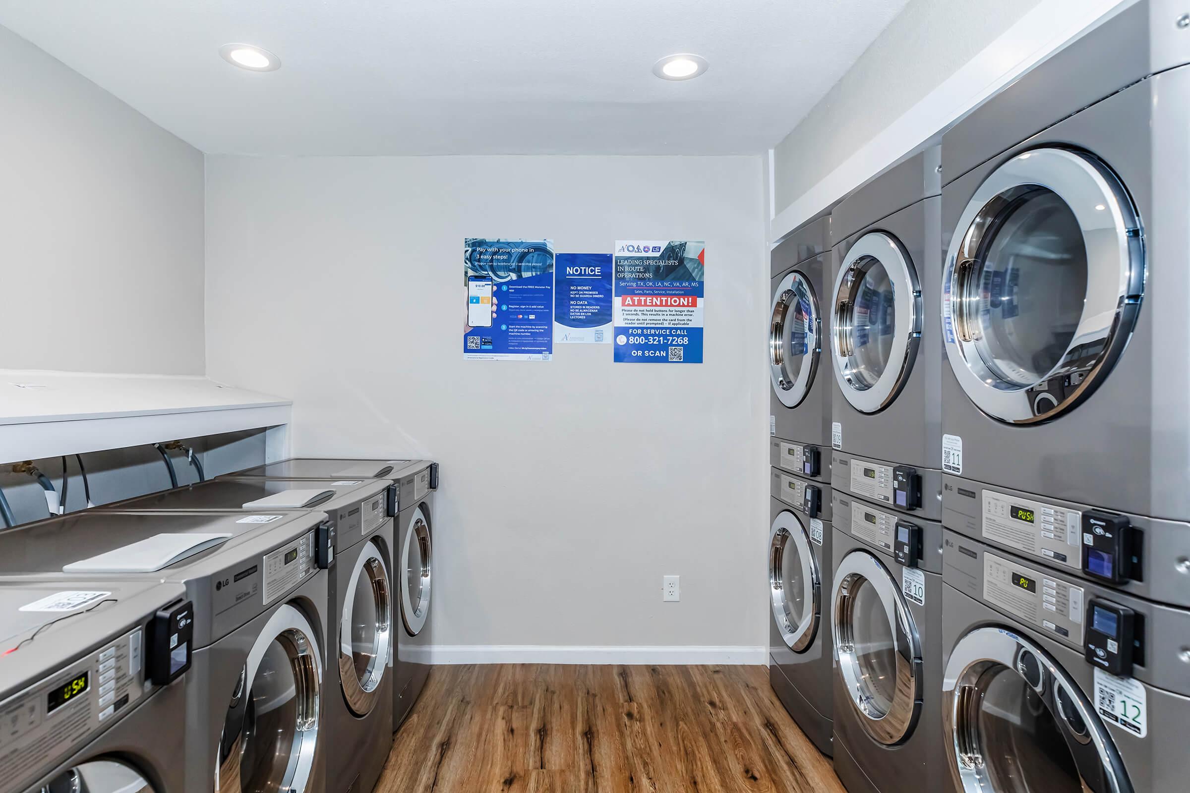 A clean laundry room featuring multiple rows of washing machines and dryers. The machines are metallic with digital displays, and there are signage and notices on the wall providing information to users. The flooring is wood-like, and the walls are painted light gray, creating a modern and tidy atmosphere.