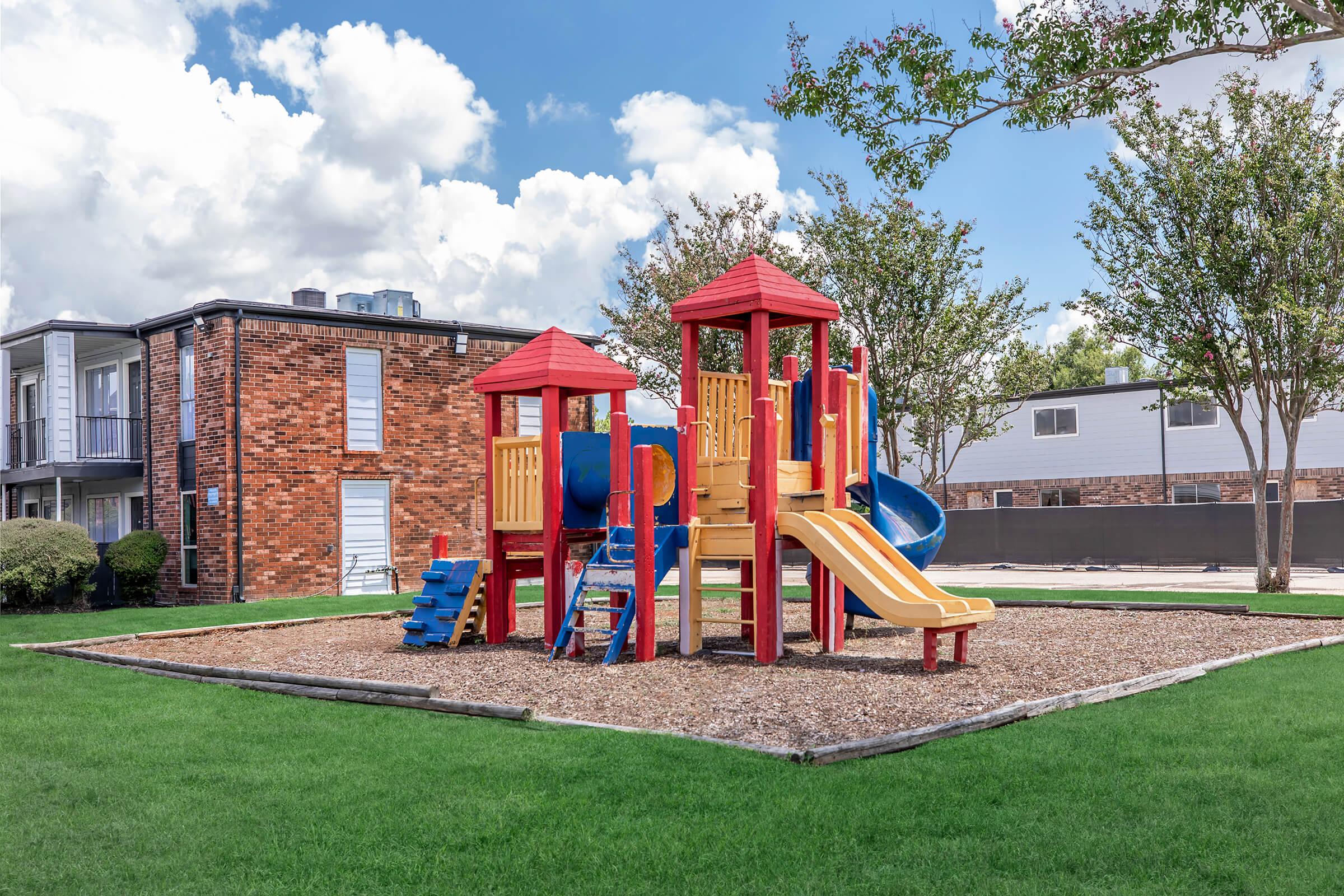 Colorful playground with a combination of slides, towers, and climbing structures, situated on a grassy area surrounded by trees and buildings. The sky is partly cloudy, enhancing the cheerful atmosphere of the play area.