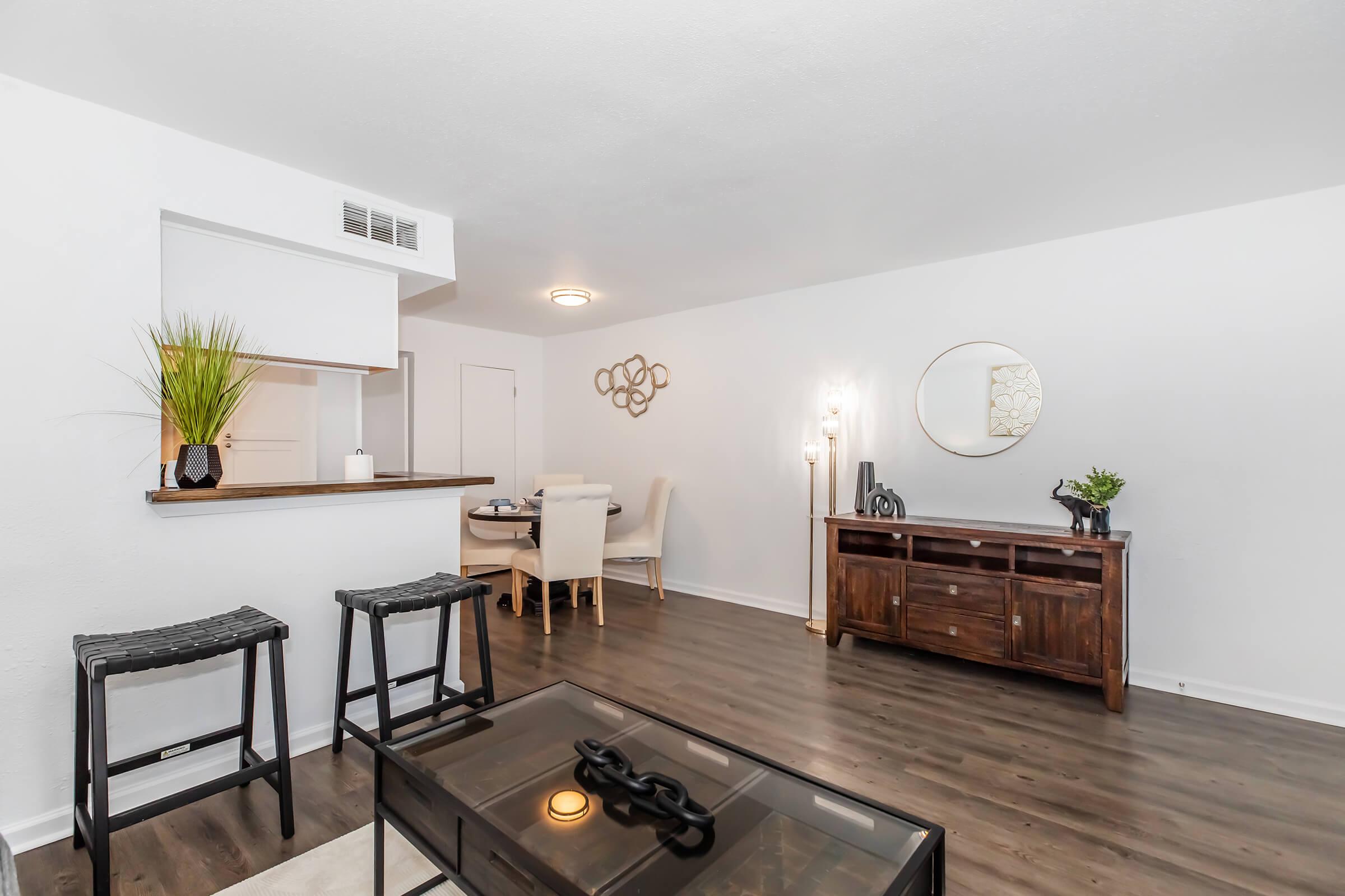 A modern living space featuring a dining area with a round table and chairs, a wooden sideboard, and a large mirror on the wall. The room has light-colored walls and dark wooden flooring. Two black stools are positioned near a glass coffee table. Decorative plants and a wall decoration enhance the ambiance.