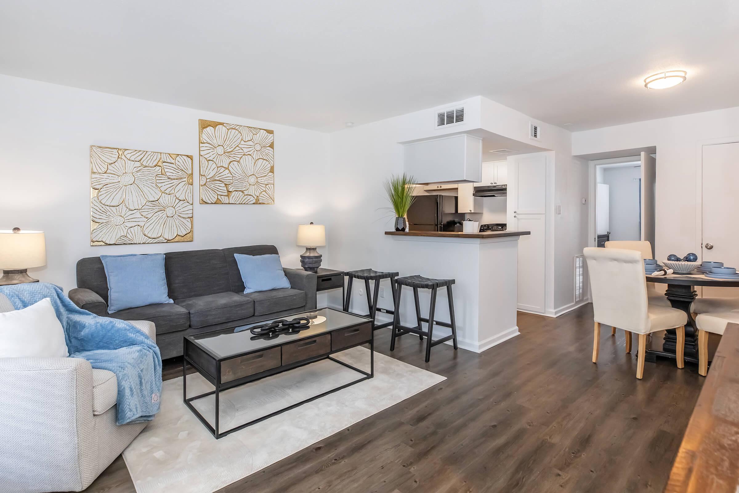 A bright and modern living space featuring a gray sofa with blue pillows, a small dining area with white chairs, and a sleek coffee table. The kitchen bar is partially visible, with decorative plants and artwork on the walls, creating a cozy and inviting atmosphere.