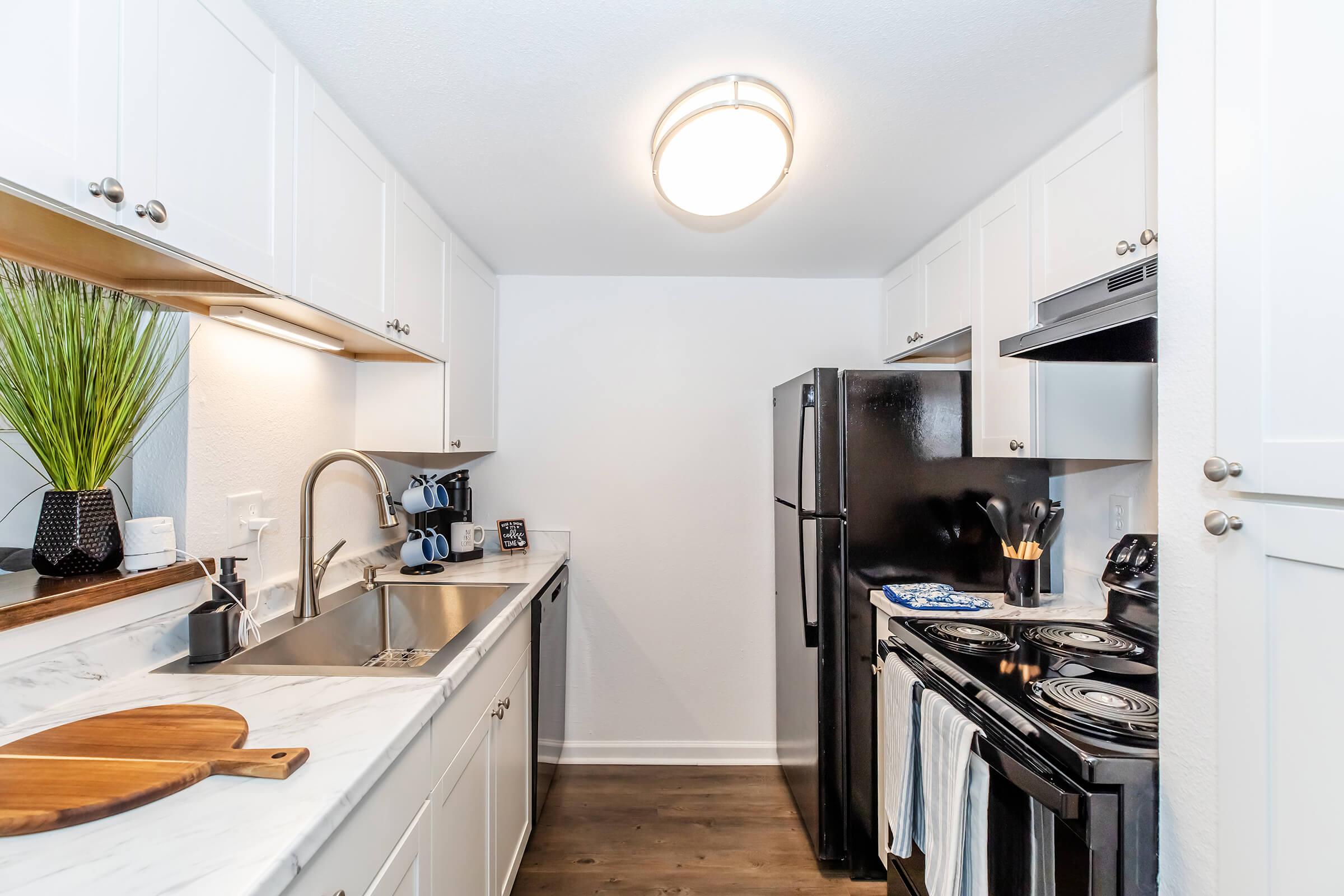 A modern kitchen with white cabinetry, a marble countertop, and stainless steel appliances. The space features a black refrigerator, a stove with oven, a sink with a faucet, and kitchen utensils. There is a wooden cutting board on the counter and a decorative plant near the window. The walls are painted light.
