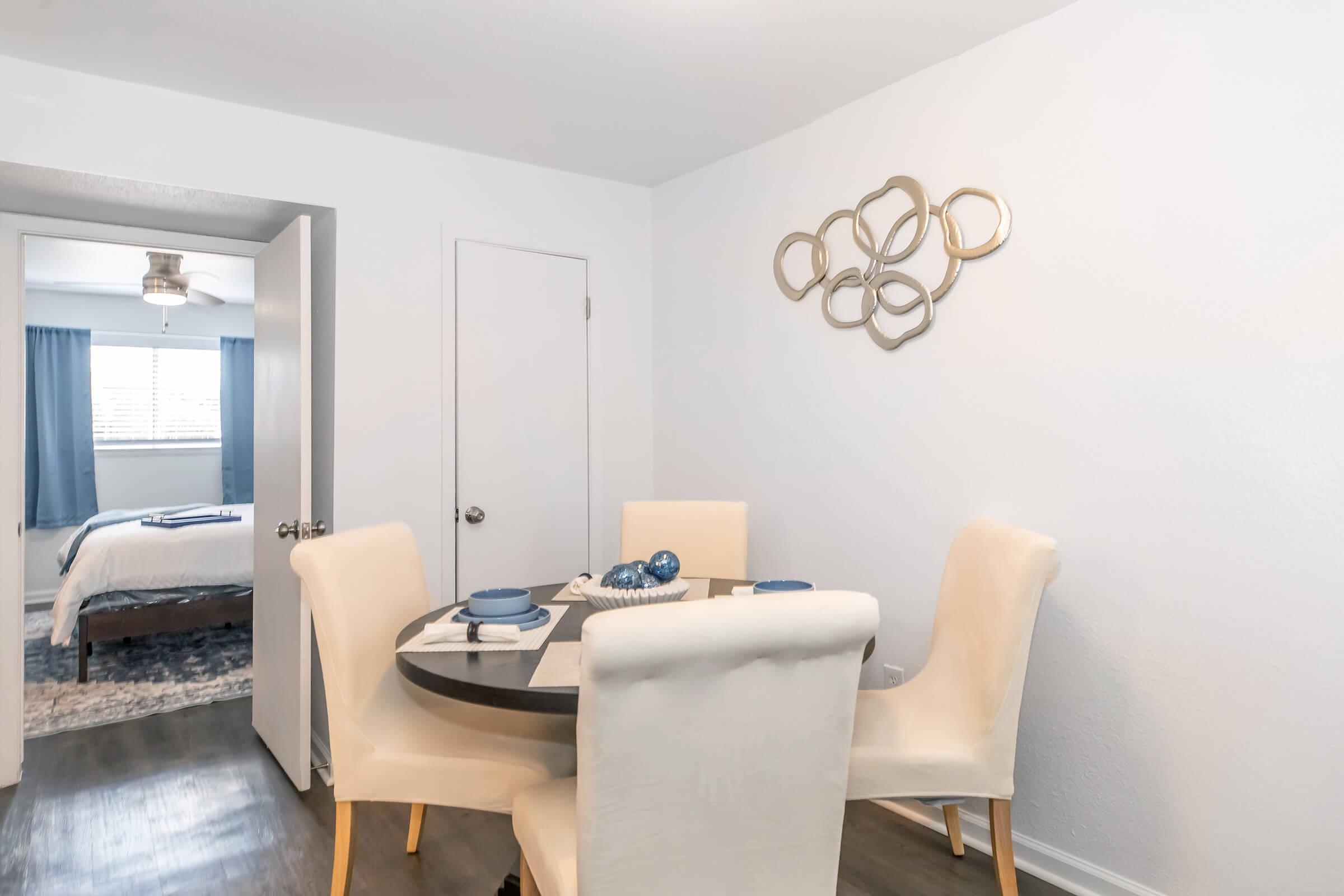 A modern dining area featuring a round table with four light-colored chairs. The table is set with blue plates and decorative items. A doorway leads to a bedroom in the background, which has a bed and blue curtains. The walls are painted white, and there is an abstract wall art piece above the table.